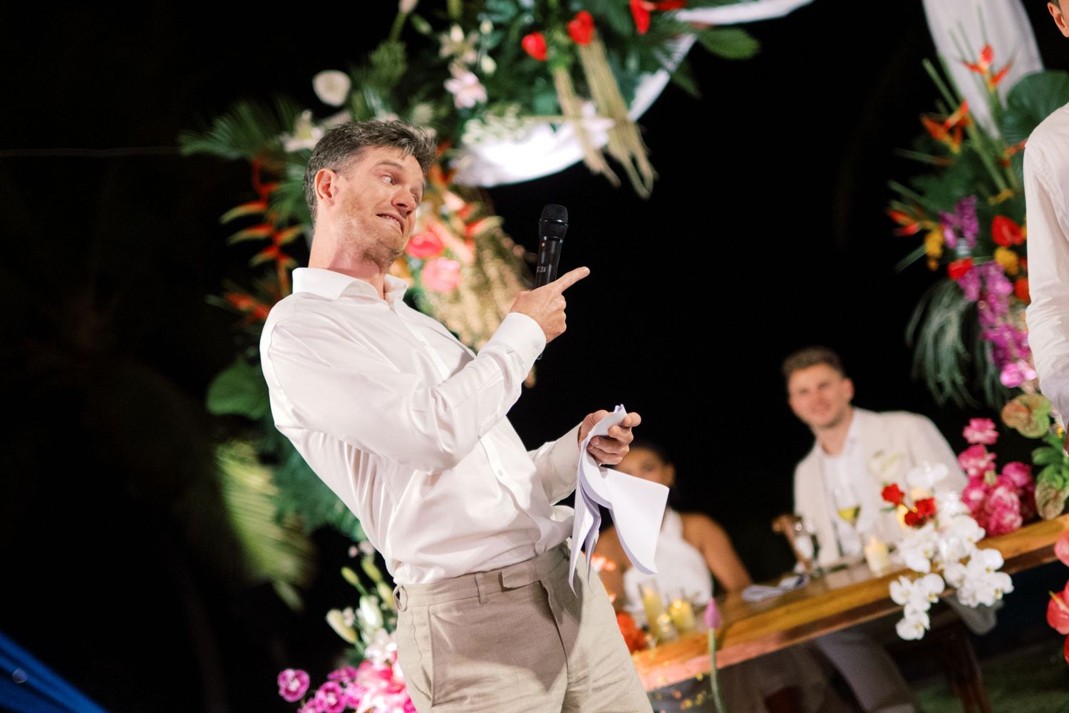 Smiling groom in white shirt gives speech at tropical floral-decorated wedding reception at night.