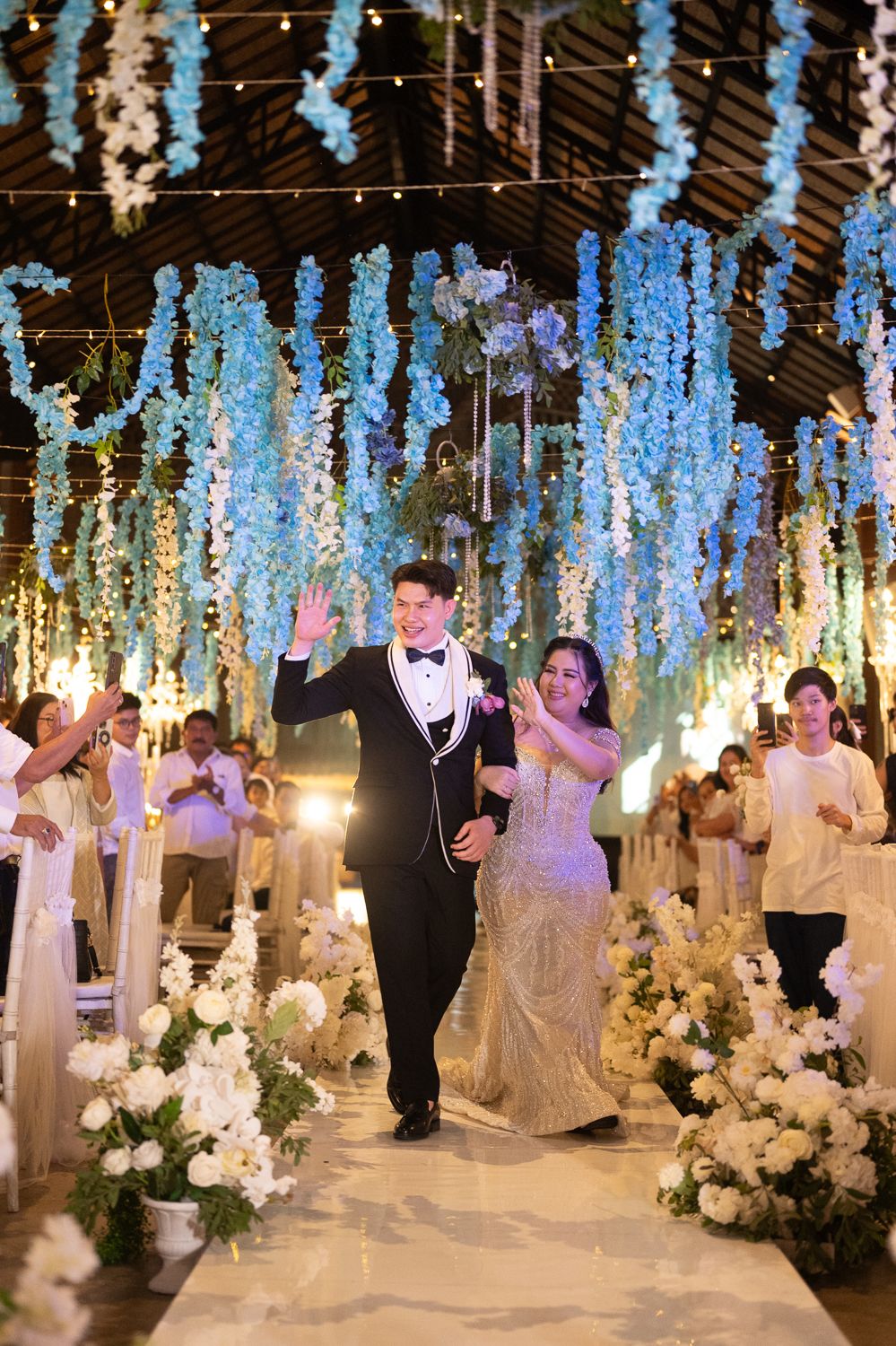 Bride and groom walk down floral aisle adorned with blue hanging flowers and white blooms at elegant wedding reception.