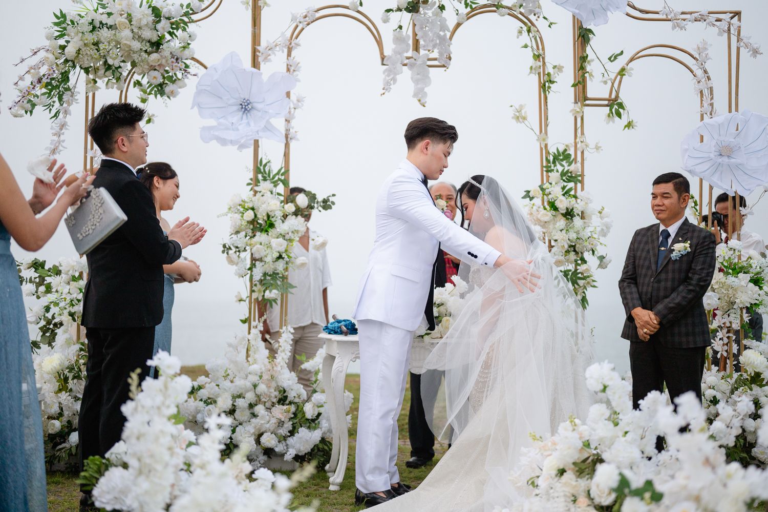 Couple exchanging vows at elegant white floral wedding ceremony with gold arch backdrop and guests applauding.