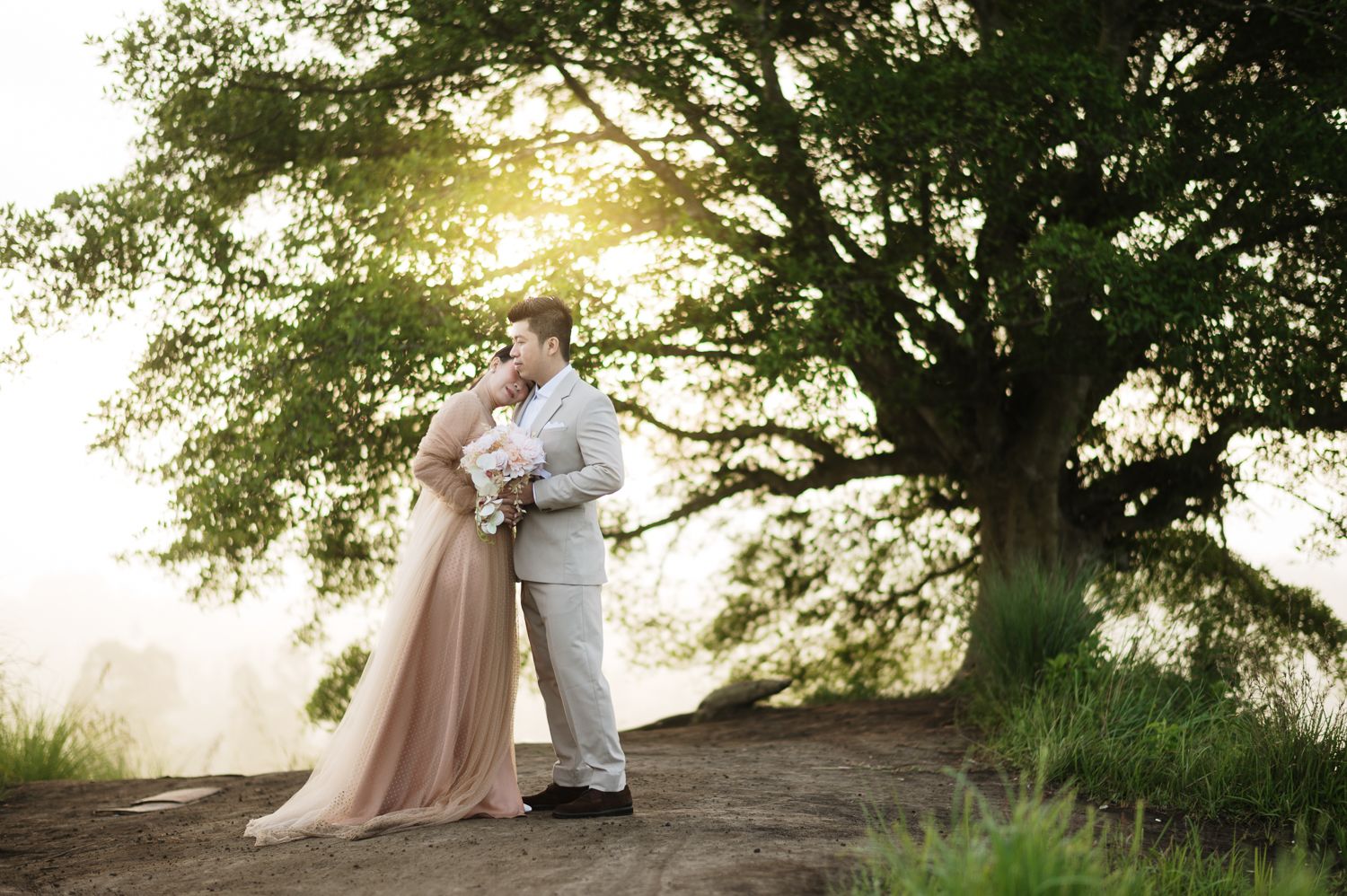 Bride and groom share romantic embrace under large oak tree at golden hour outdoor wedding ceremony.