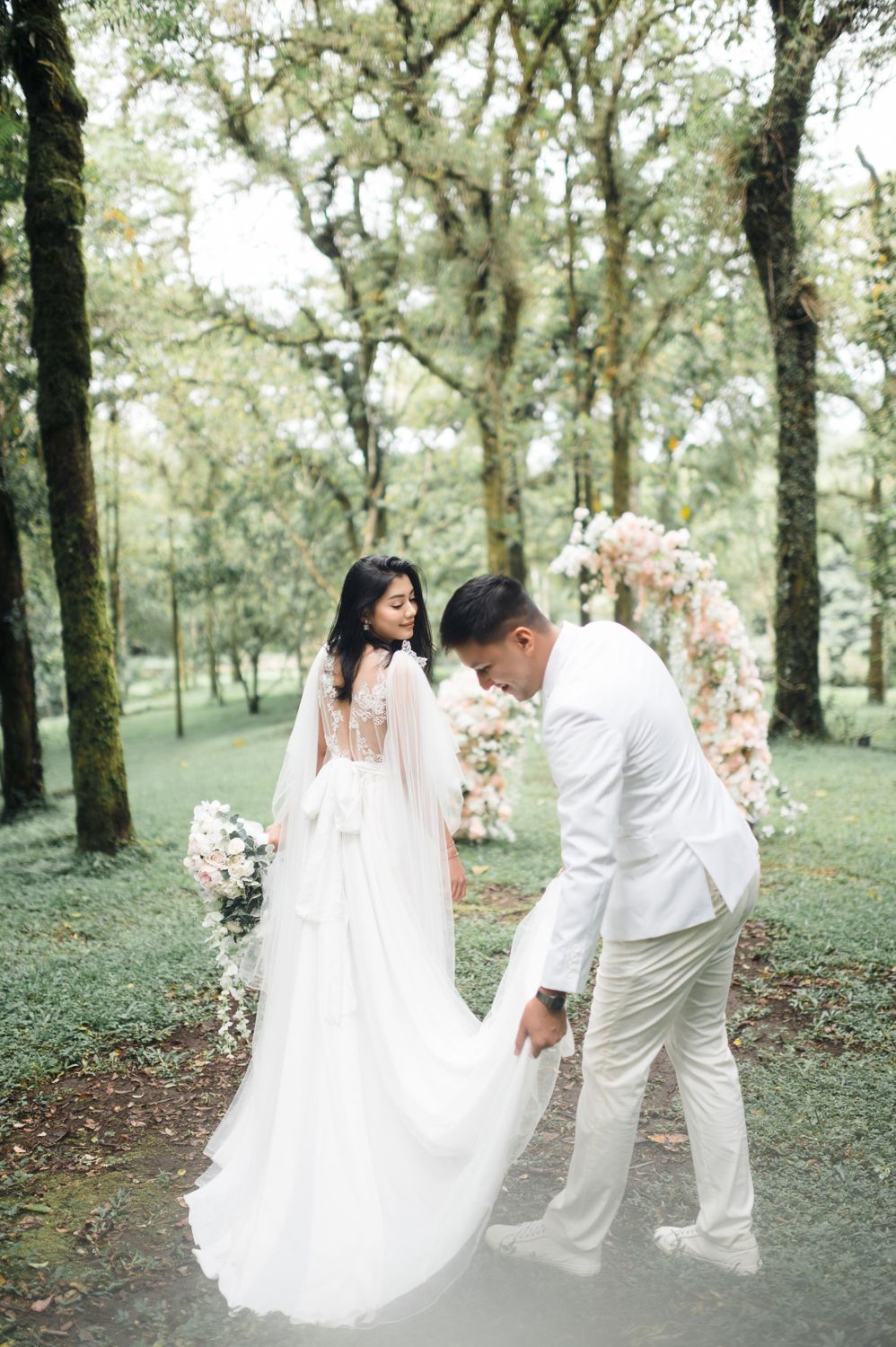 Groom in white suit adjusting bride's flowing wedding gown train in a lush green forest ceremony setting.