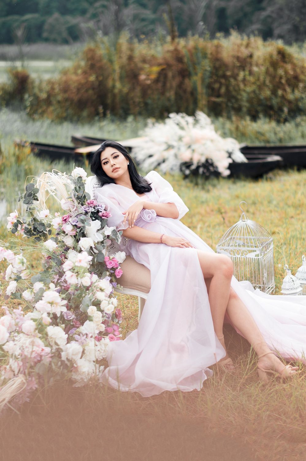 A woman in a flowing pink dress sits gracefully among white flowers near a lake with boats in the background.