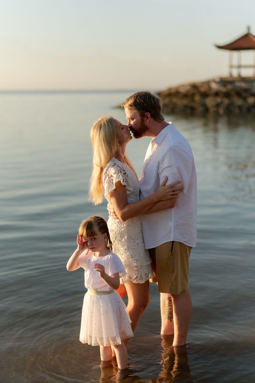 Couple sharing a romantic kiss while standing in shallow water at beach sunset with child nearby.