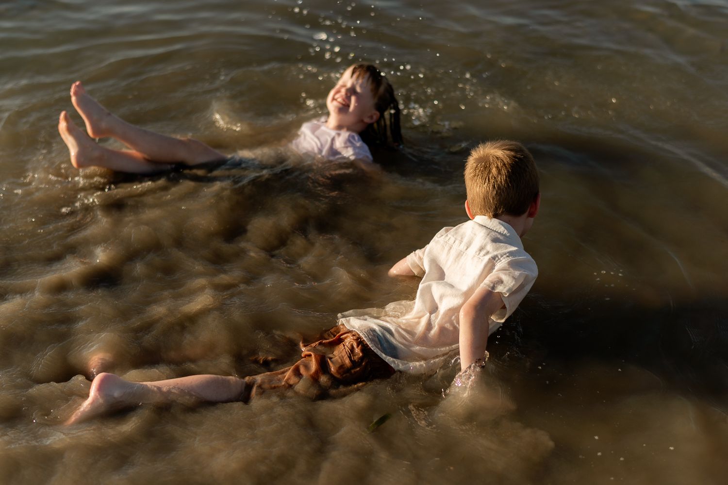 Two children play and splash in shallow sunlit ocean water near the shore.