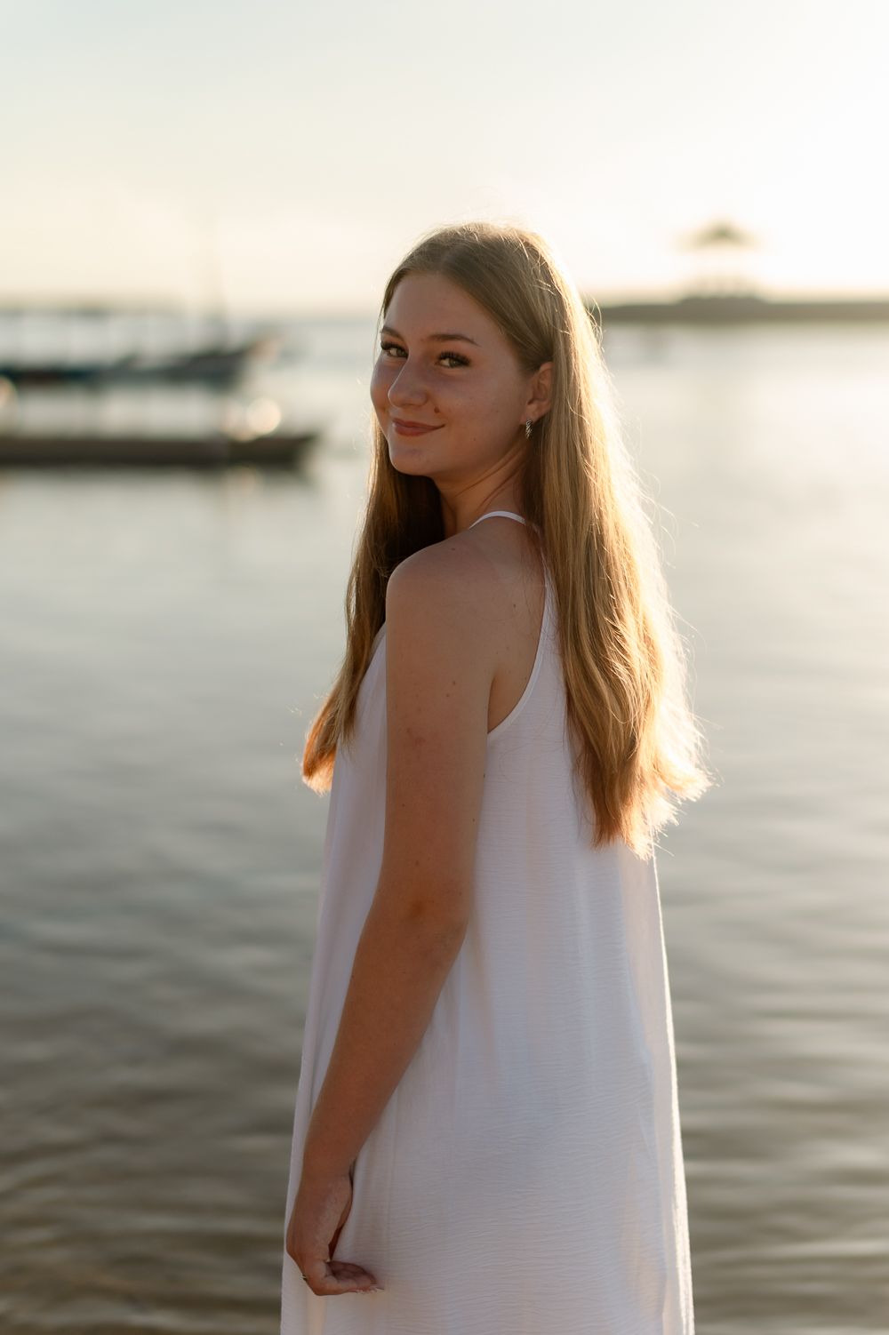 Young blonde woman in white sundress smiles over shoulder at golden hour beach.