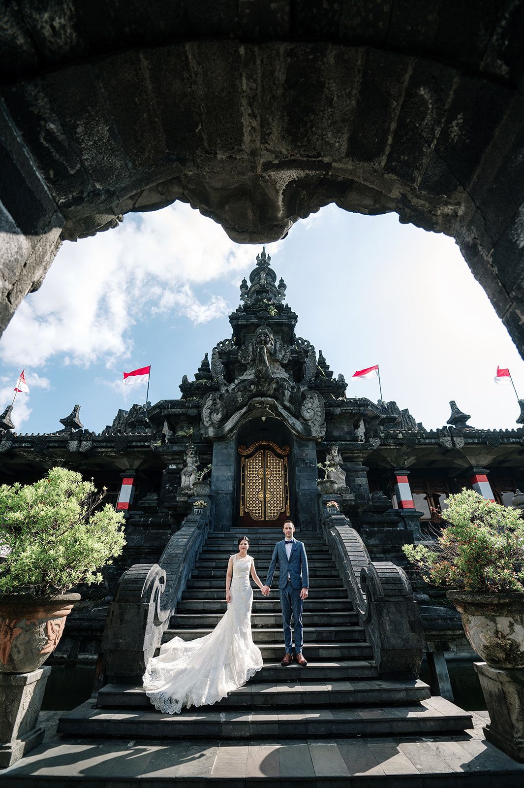 Couple in wedding attire poses on ornate Balinese temple steps with red flags and intricate stone carvings.