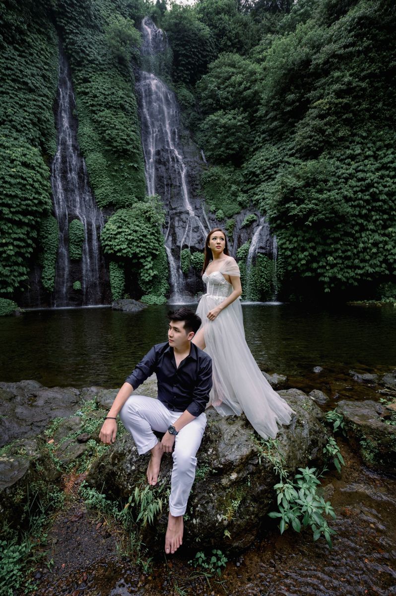Couple posing by a lush waterfall, woman in white gown standing on rocks, man seated beside her.