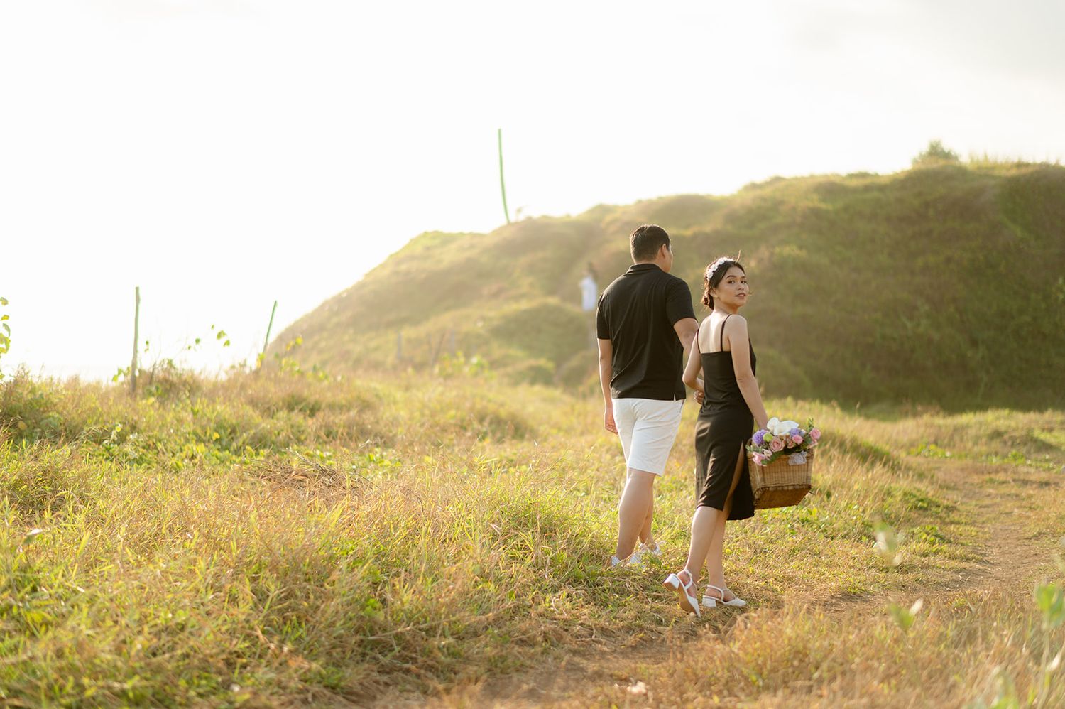 Couple walking along a scenic grassy hillside path at golden hour, woman carrying a picnic basket.
