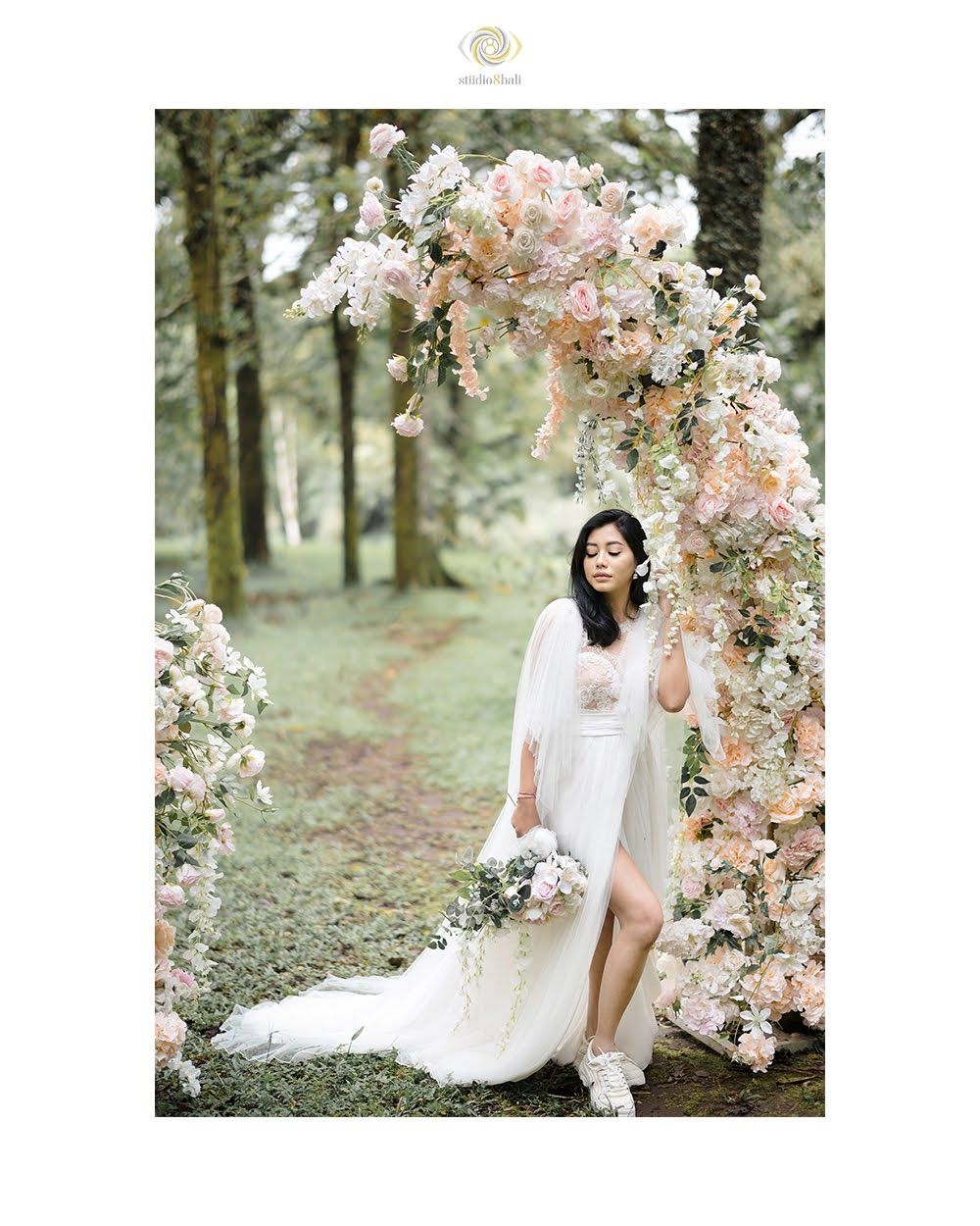 Bride in white gown poses under floral arch with pink roses in a lush green forest setting.