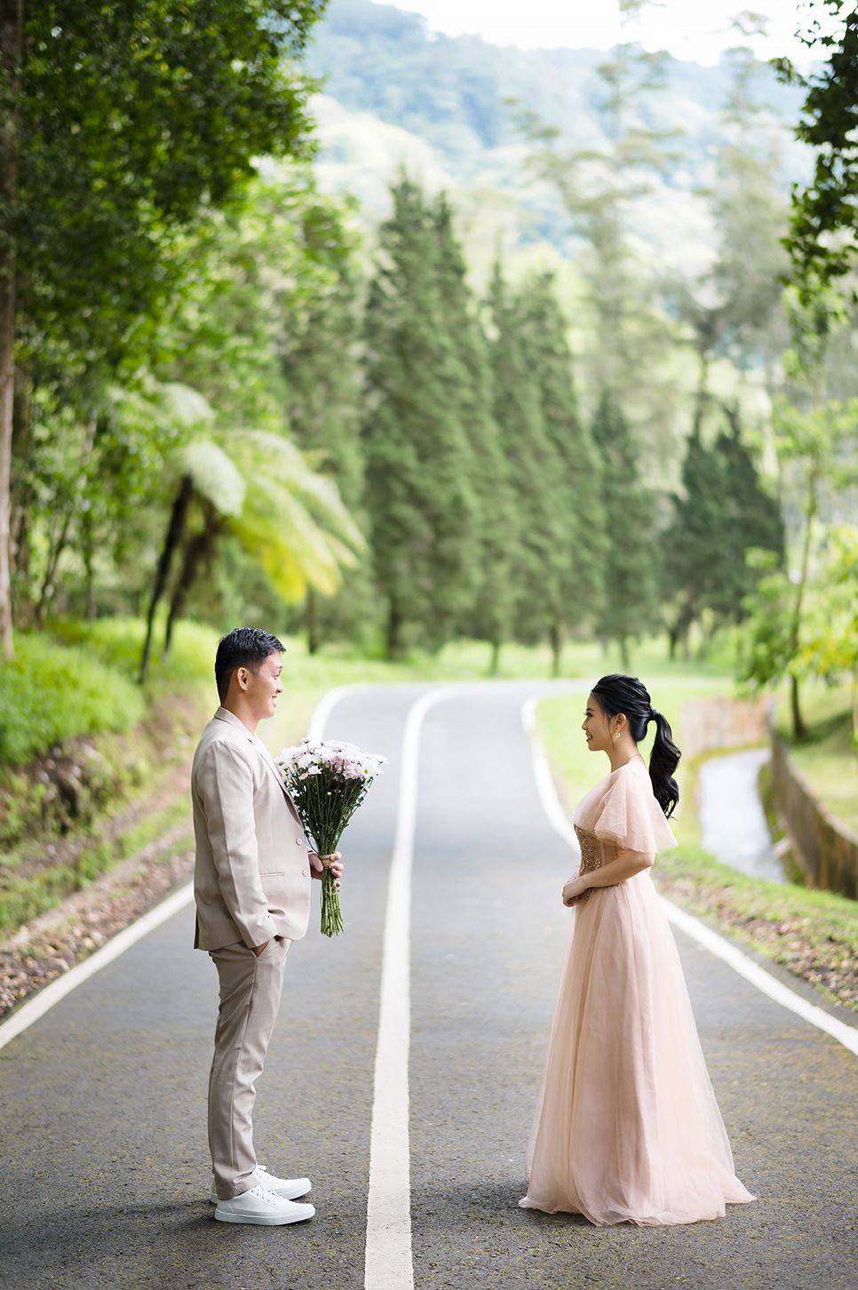 Man presenting flowers to woman in pink gown on a scenic tree-lined road during an outdoor pre-wedding shoot.