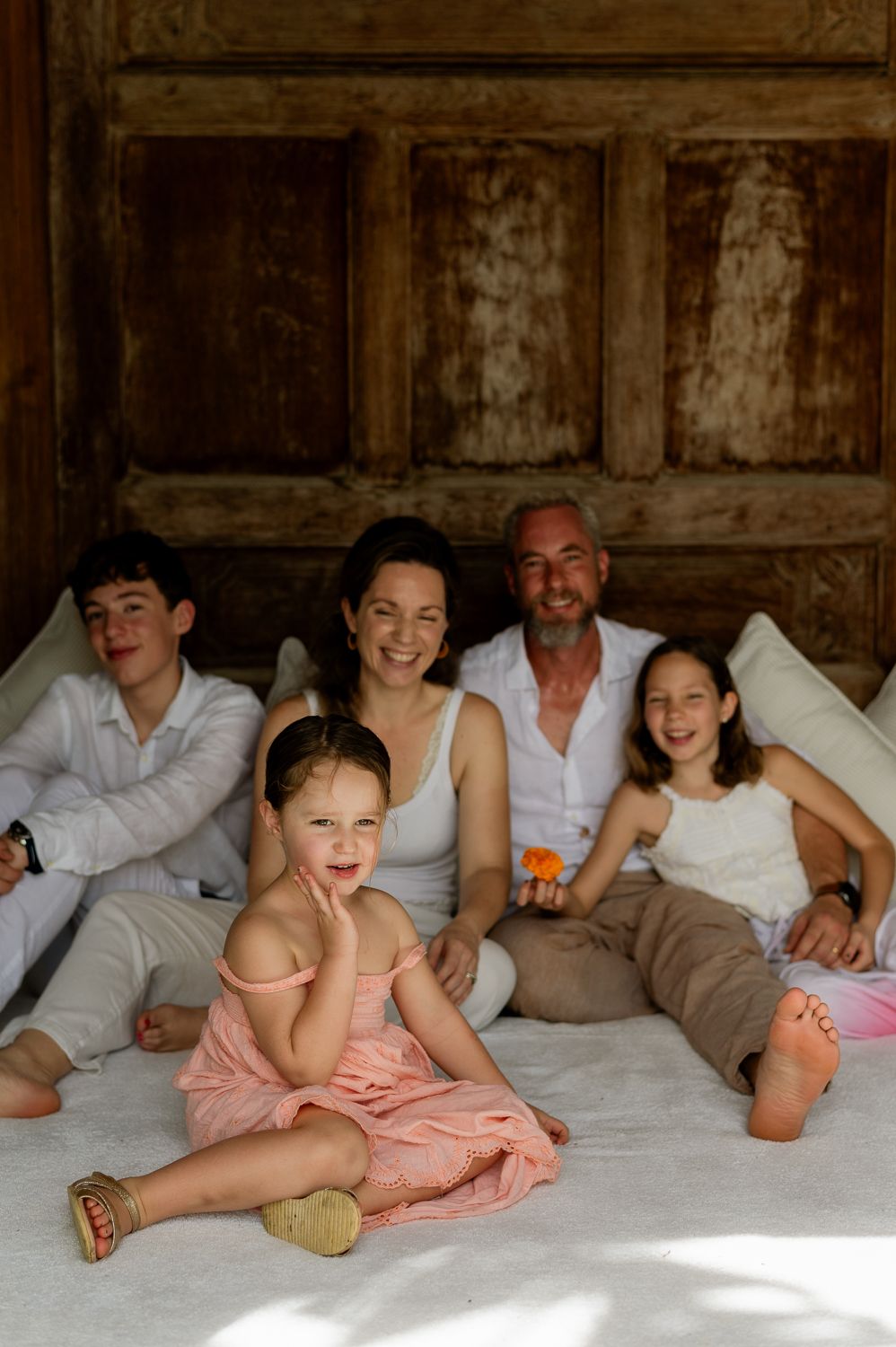 Happy family of five posing together on a white bed, smiling, with rustic wooden door backdrop.