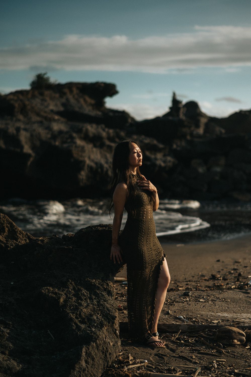 Woman in olive crochet dress poses on rocky beach at golden hour, waves and dramatic rock formations behind her.