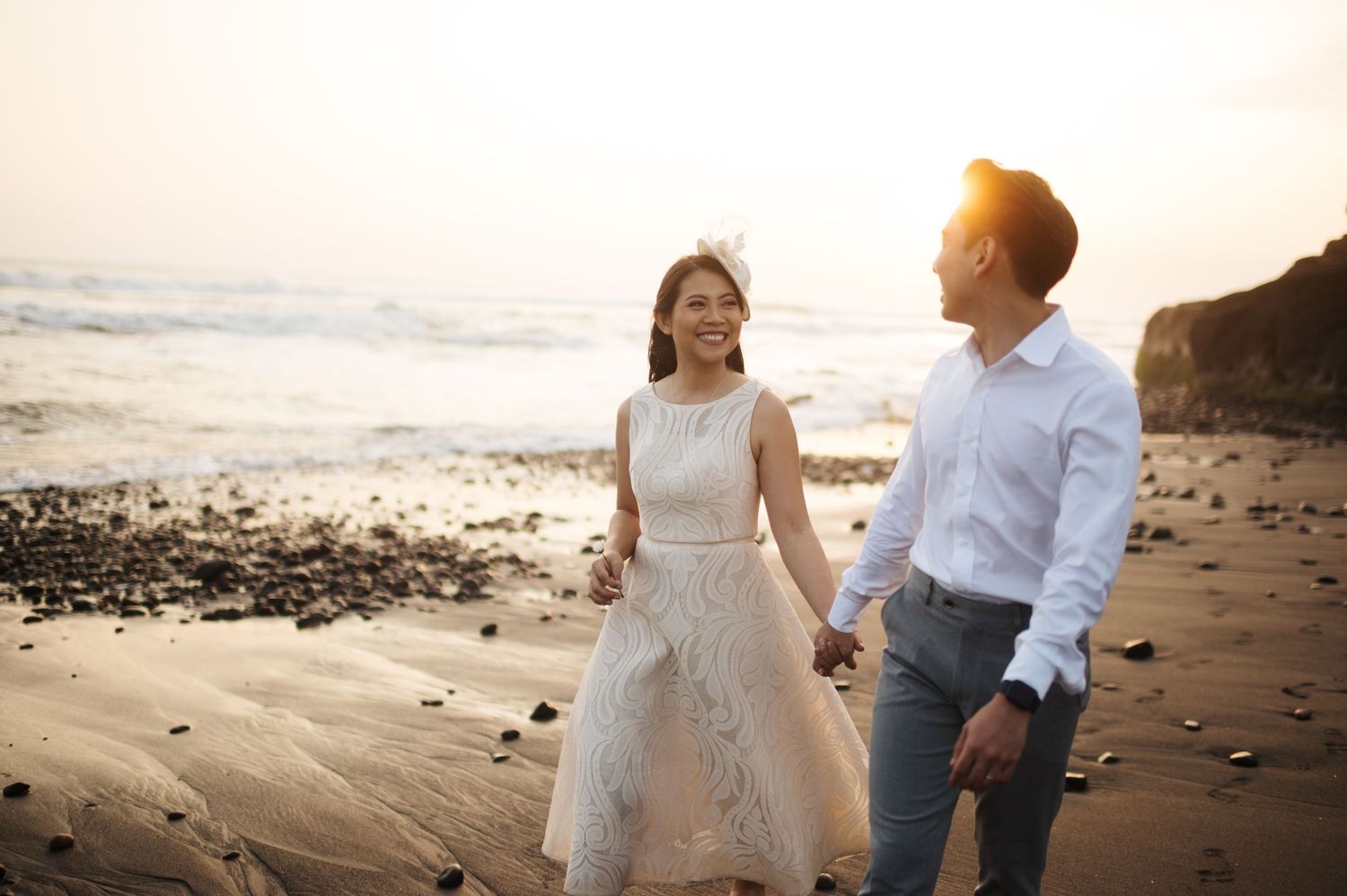 Couple holding hands walking on a sandy beach at sunset, woman in white dress, man in white shirt.
