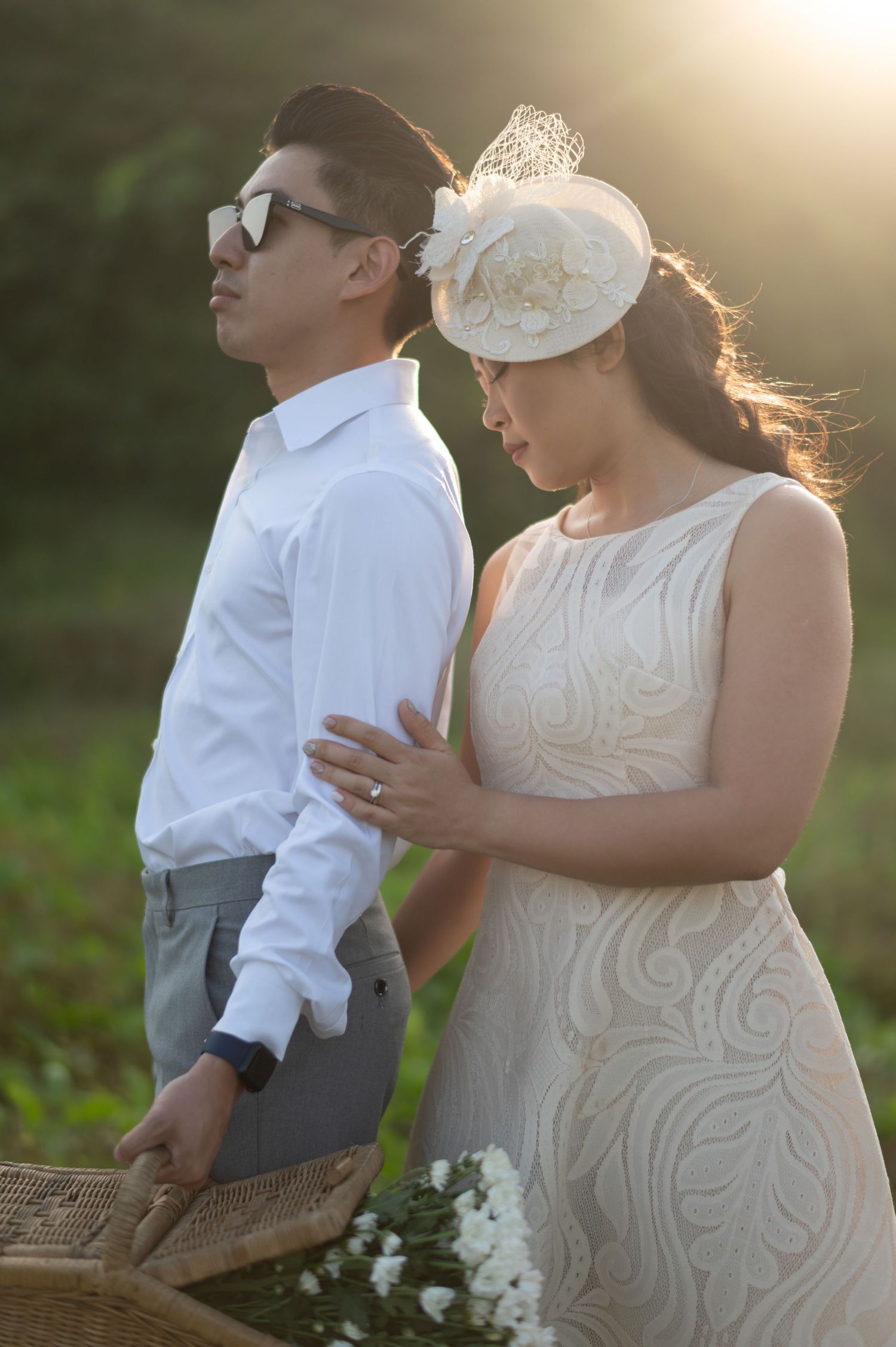 Couple in elegant wedding attire poses outdoors at golden hour, woman in lace dress and fascinator holds groom's arm.