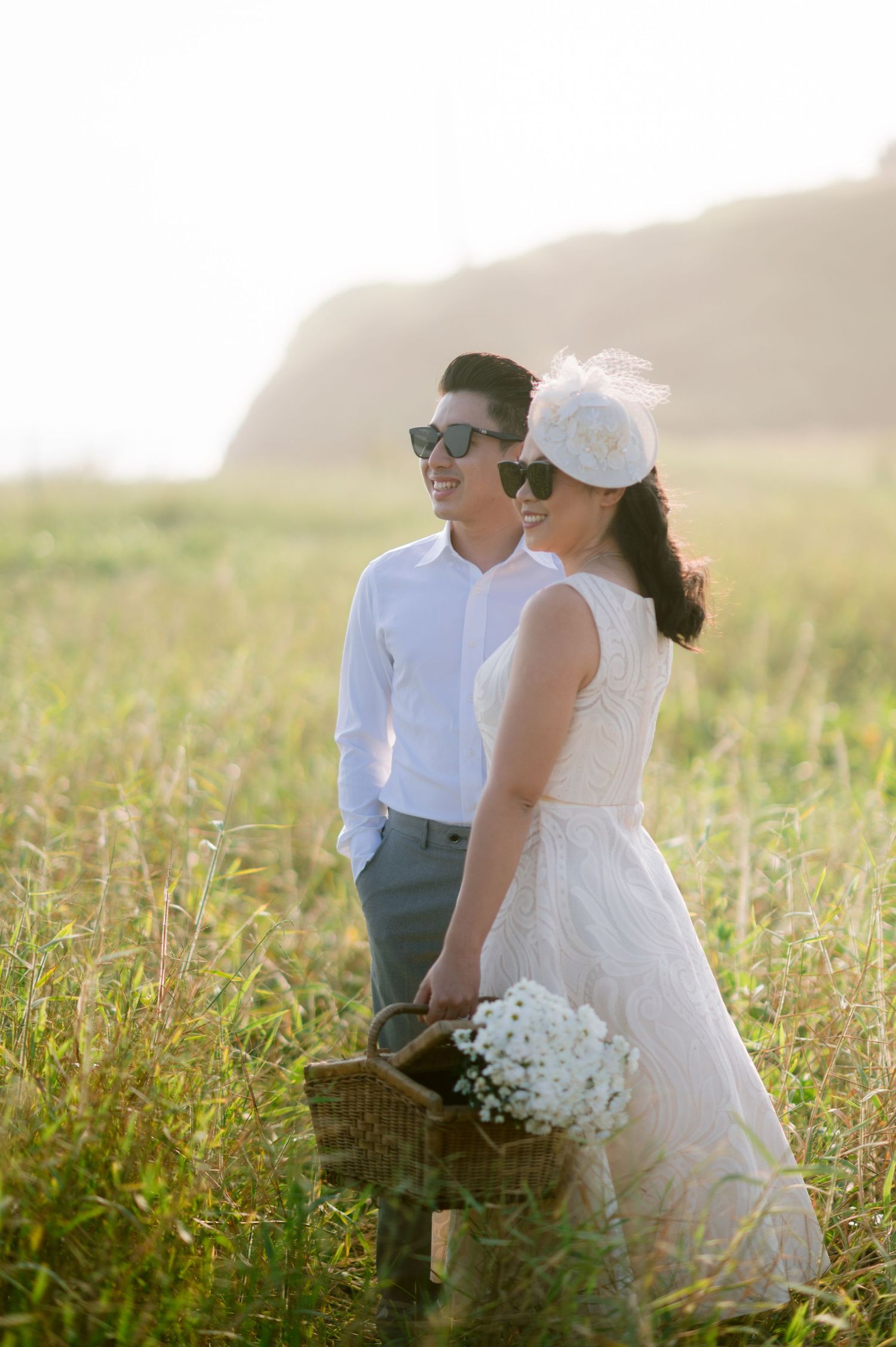 Couple in wedding attire wearing sunglasses standing in a golden field, bride holding a wicker basket with white flowers.