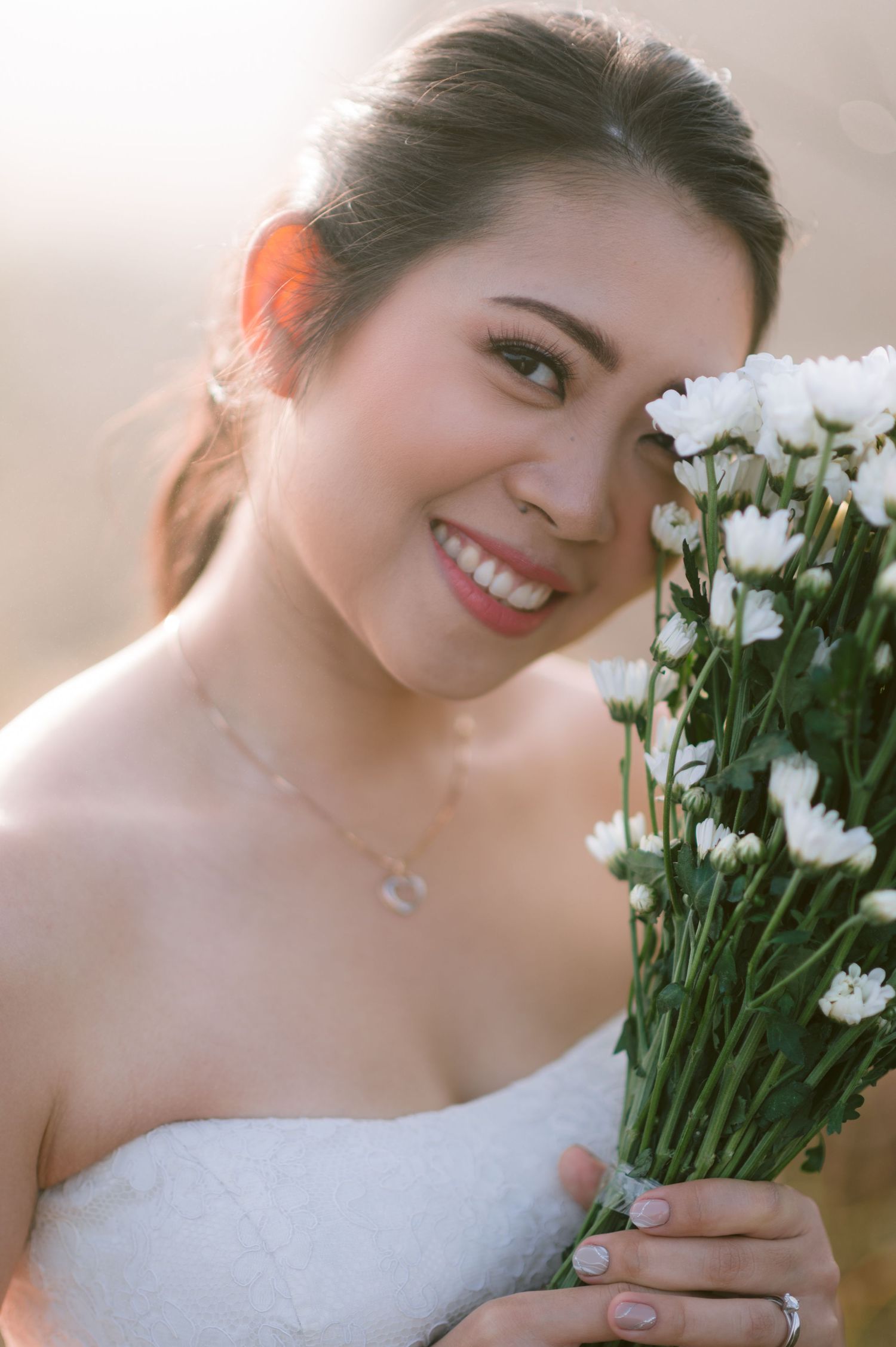 Smiling bride in white dress holding bouquet of white flowers, with soft golden backlight and elegant necklace.