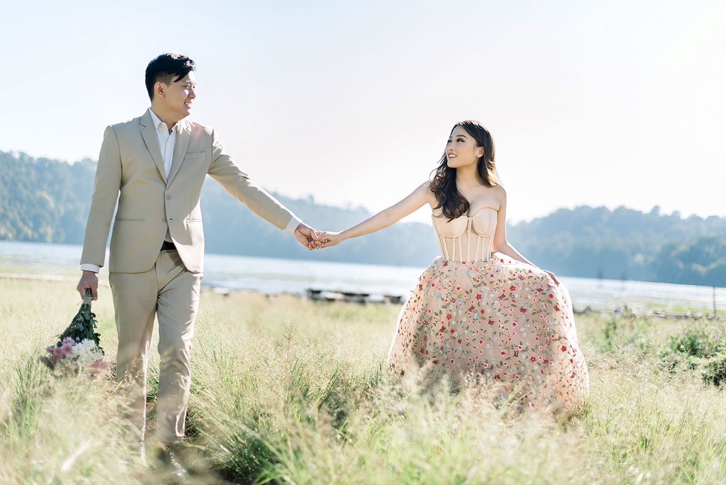 Couple holding hands in a field, man in beige suit, woman in floral pink gown, with a serene lake backdrop.
