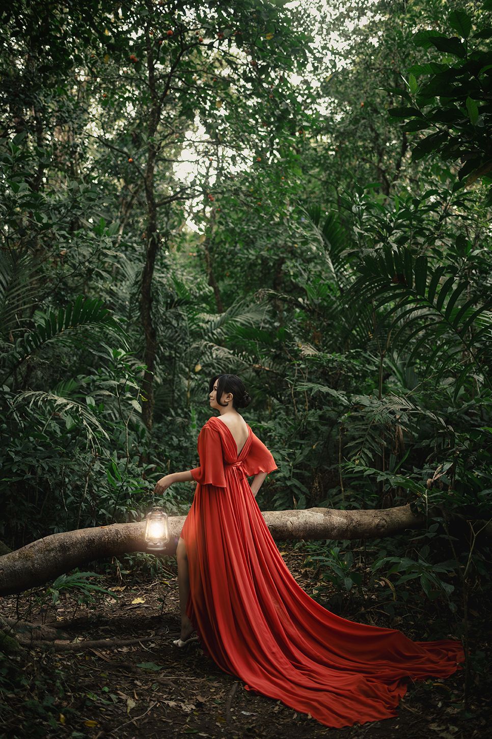 Woman in red gown holding lantern in lush forest.