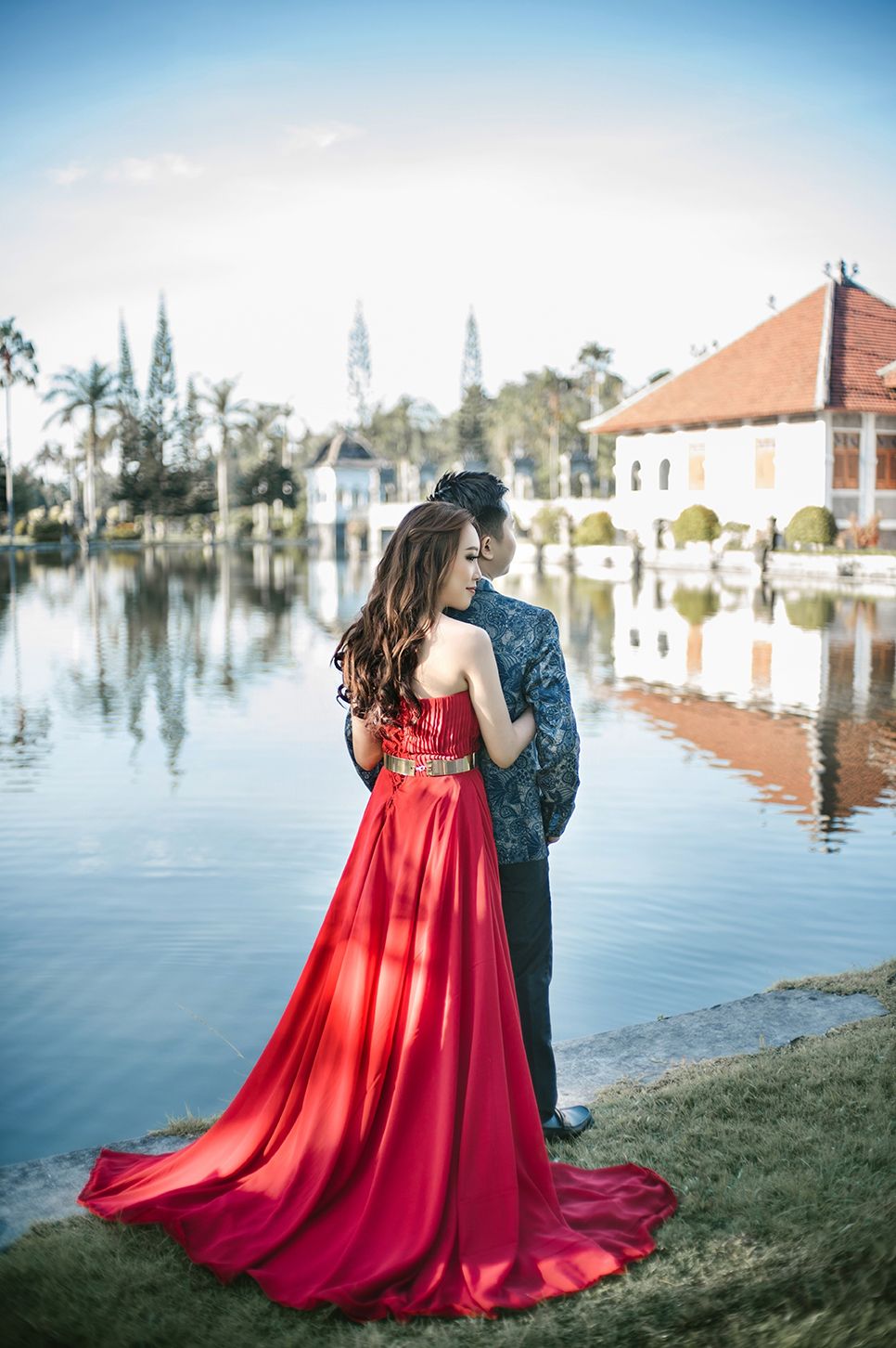 Couple in elegant attire poses by a serene lake, woman in red gown, man in dark suit, with buildings reflected in water.
