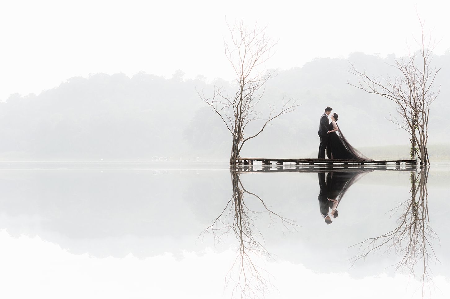 Couple shares romantic kiss on misty lake dock with bare trees and perfect water reflection in dreamy white landscape.