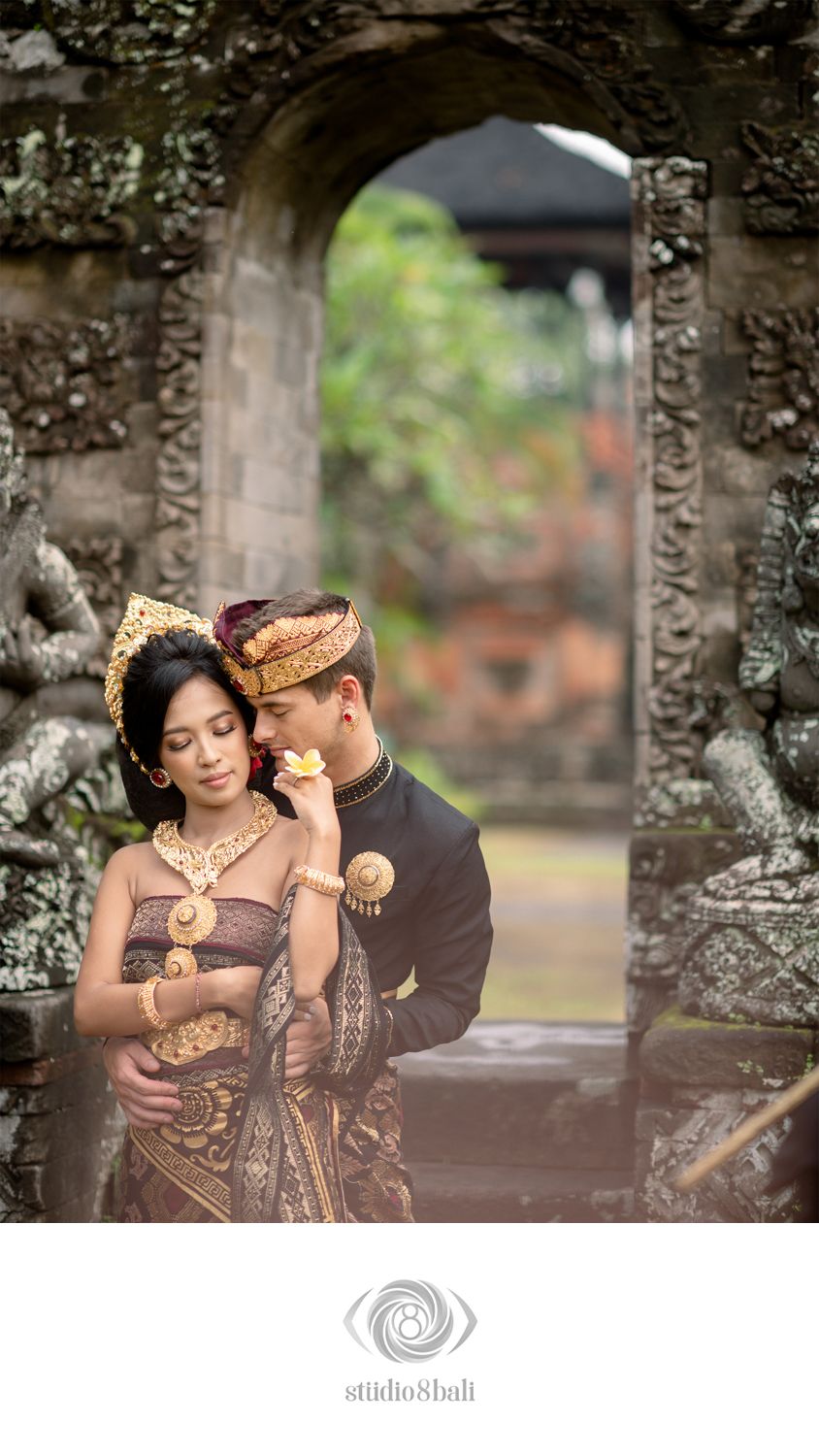 Balinese couple in traditional gold ceremonial attire posing romantically in an ornate stone temple gateway in Bali.