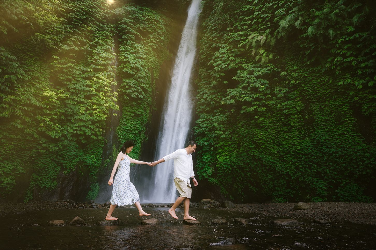 Couple in white holding hands walking near a lush green waterfall in a romantic outdoor setting.