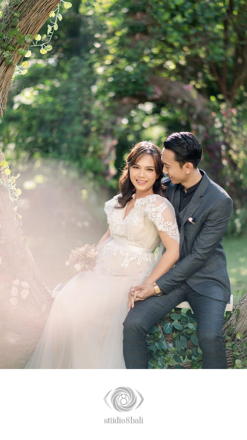 Couple in wedding attire sitting together in a lush garden, bride in lace gown holding bouquet, groom in gray suit.