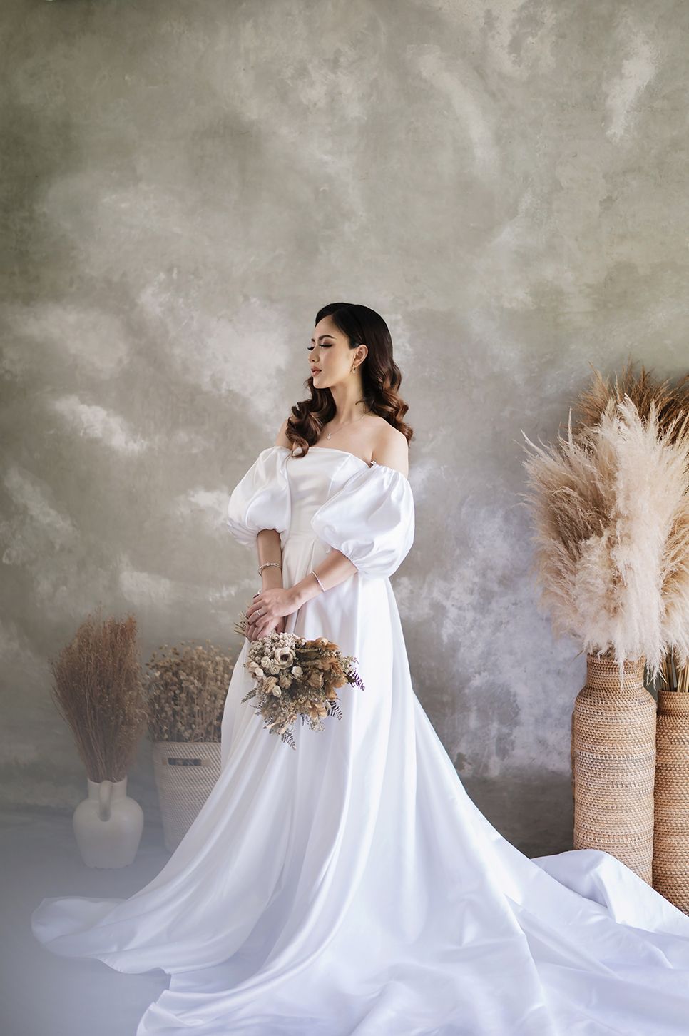 Bride in white off-shoulder gown with puff sleeves holds bouquet, posing before textured wall with pampas grass decor.