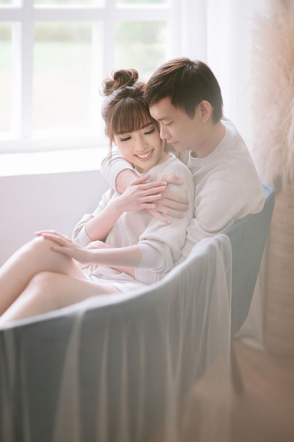 A couple shares a tender embrace in a softly lit room, with the man holding the smiling woman from behind in a chair.