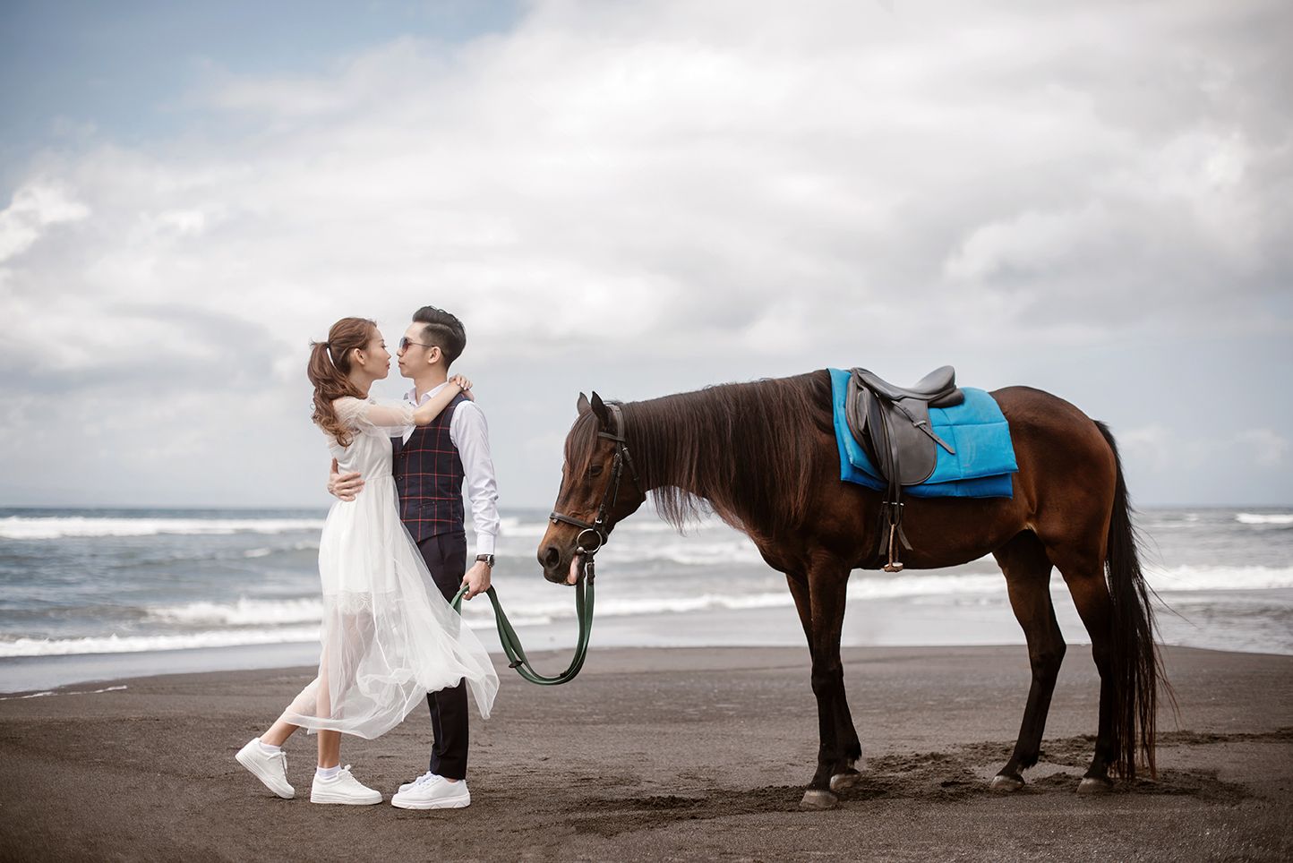 Bride and groom share a romantic moment beside a brown horse with blue saddle on a black sand beach.
