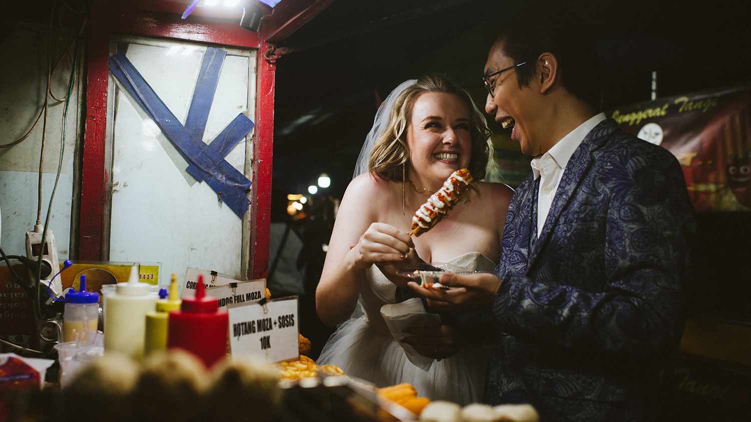 Bride and groom laughing at a food stall at night, sharing a snack in wedding attire at an outdoor market.