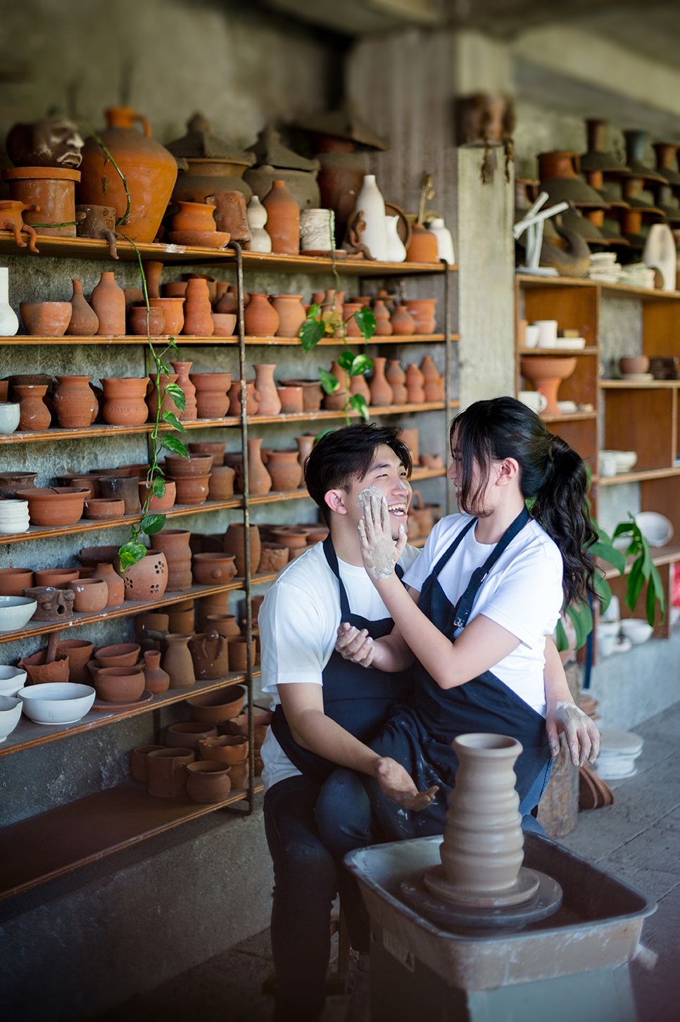 Couple in aprons shares a romantic moment at a pottery wheel surrounded by clay pots on shelves in a ceramic studio.