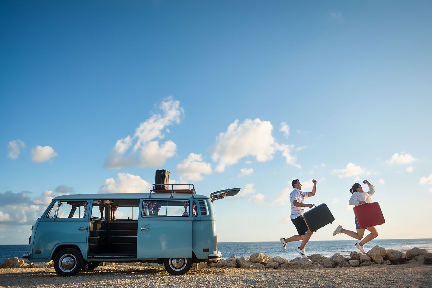 Couple jumping joyfully beside a vintage blue VW van on a sunny coastal road trip.