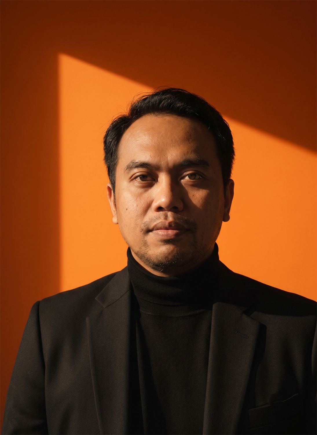 Man in black turtleneck and blazer posing against vibrant orange wall with dramatic light shadow.
