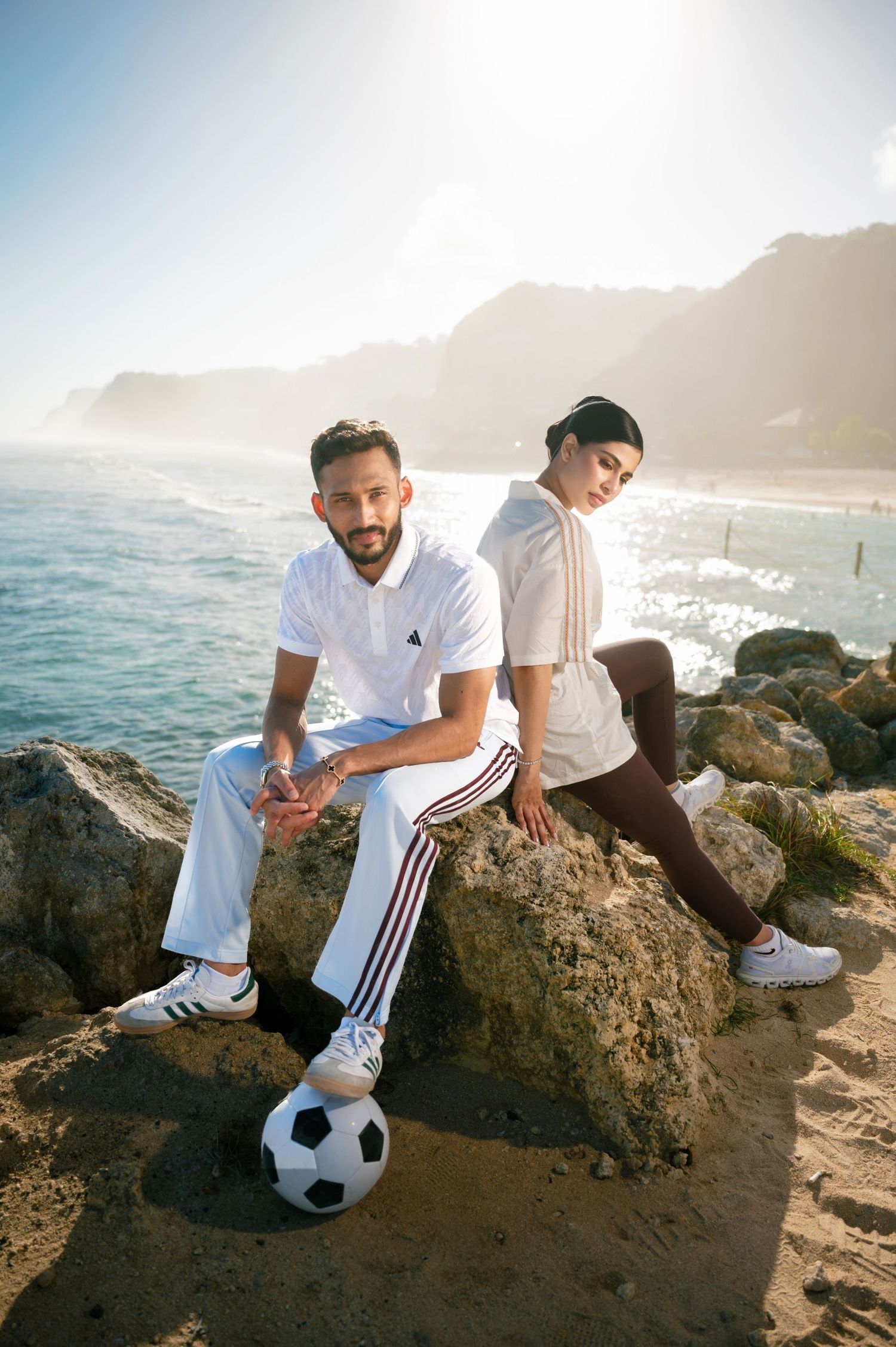 Man and woman in white Adidas sportswear sitting on rocks by the beach with a soccer ball.
