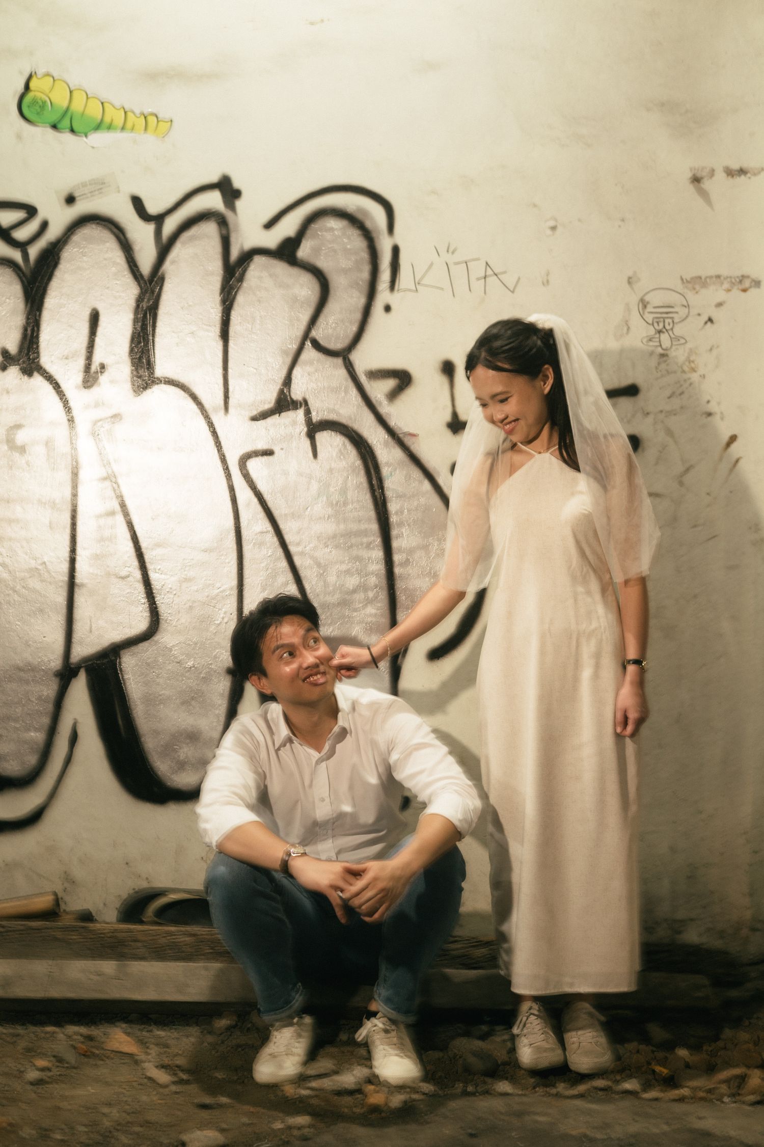 A couple poses in front of a graffiti wall, the bride in a white dress and veil, groom sitting casually below her.