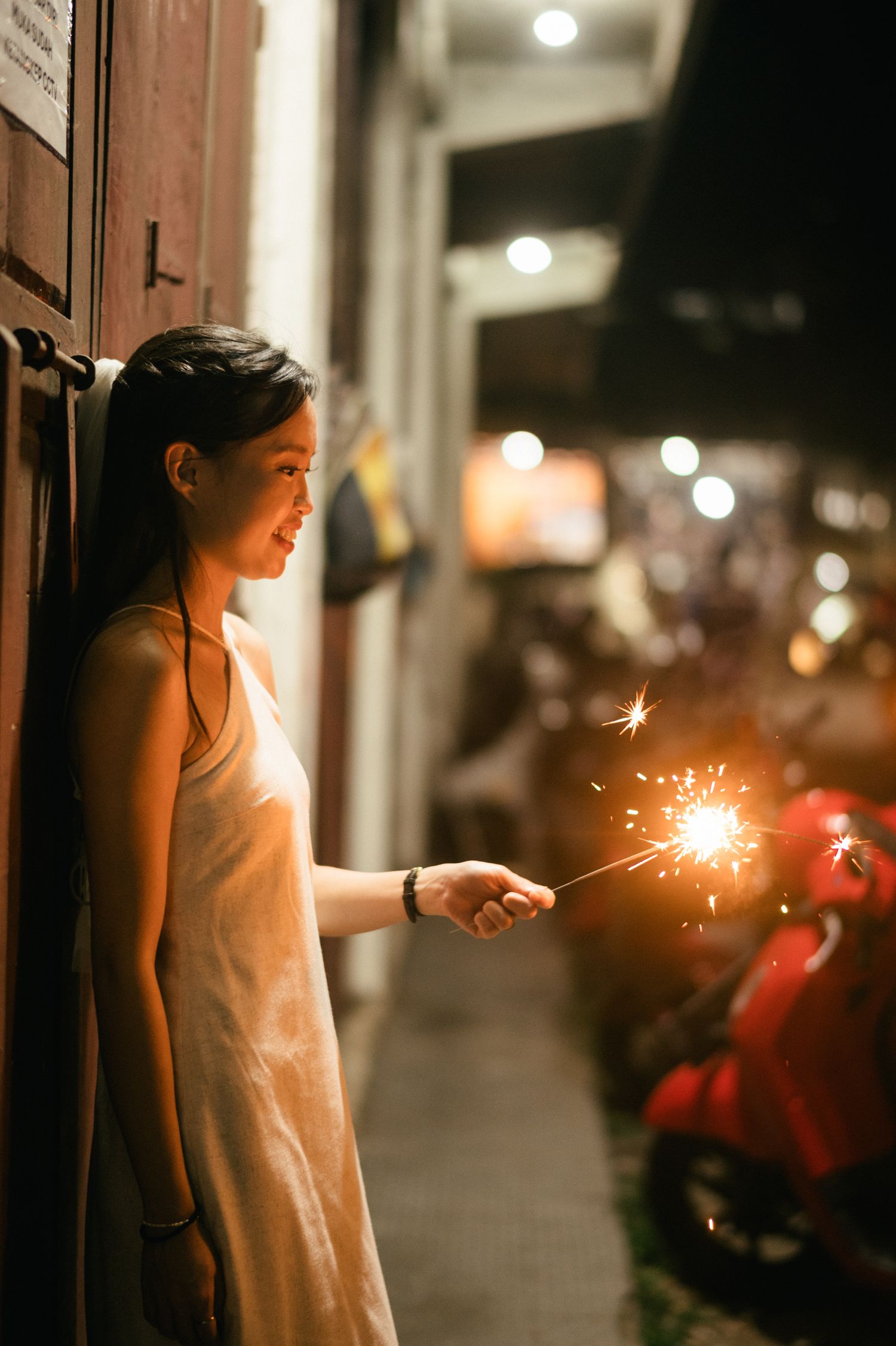 Young Asian woman in a white dress holding a sparkler at night, smiling beside a wooden door.