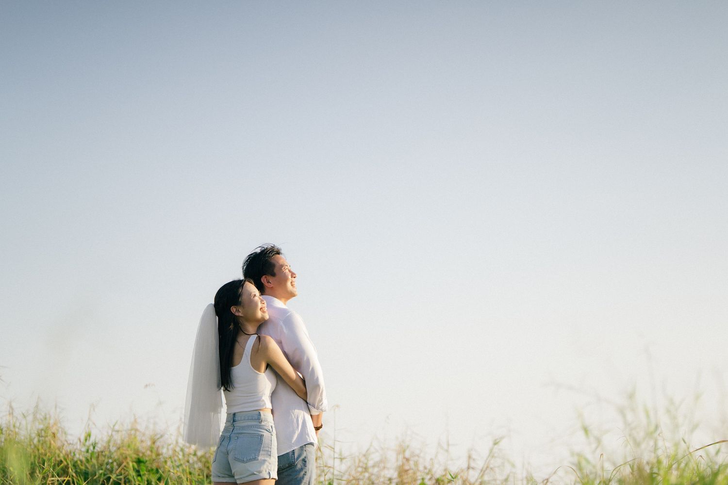 Romantic couple standing in a meadow, bride in white veil embracing groom under a clear sky.