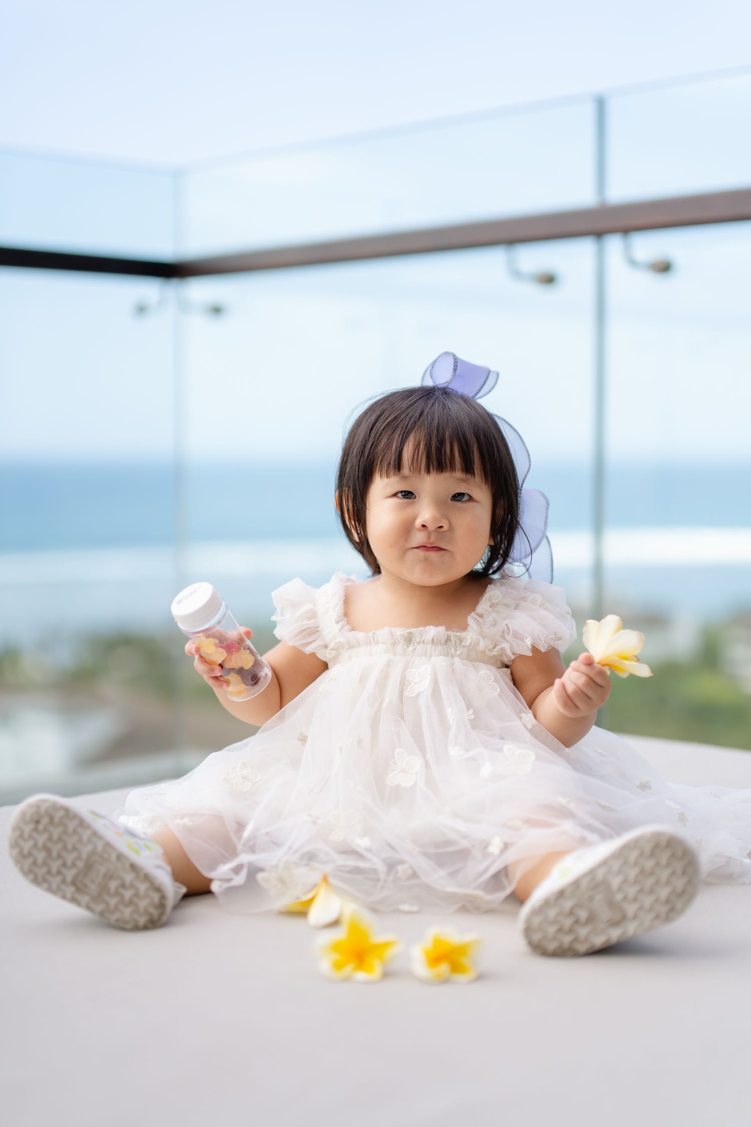 A cute toddler girl in a white lace dress with a purple bow sits on a floor holding flowers and a bottle.