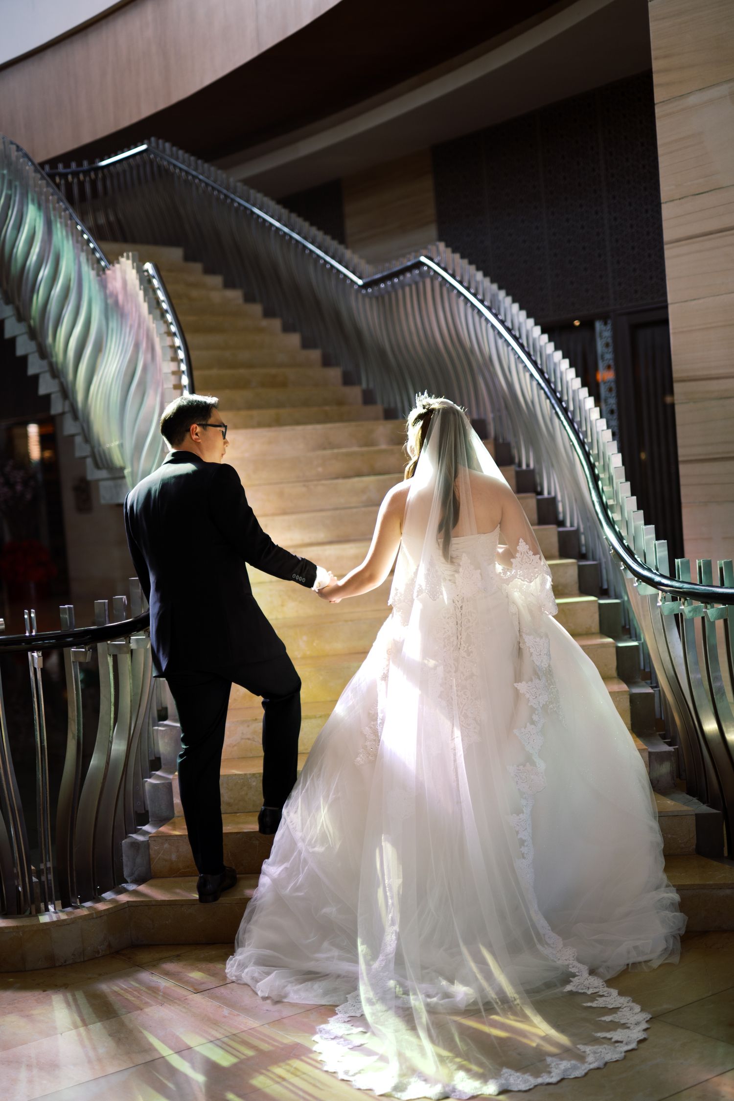 Bride and groom holding hands walking up elegant staircase, bride in white lace gown with veil, groom in black tuxedo.