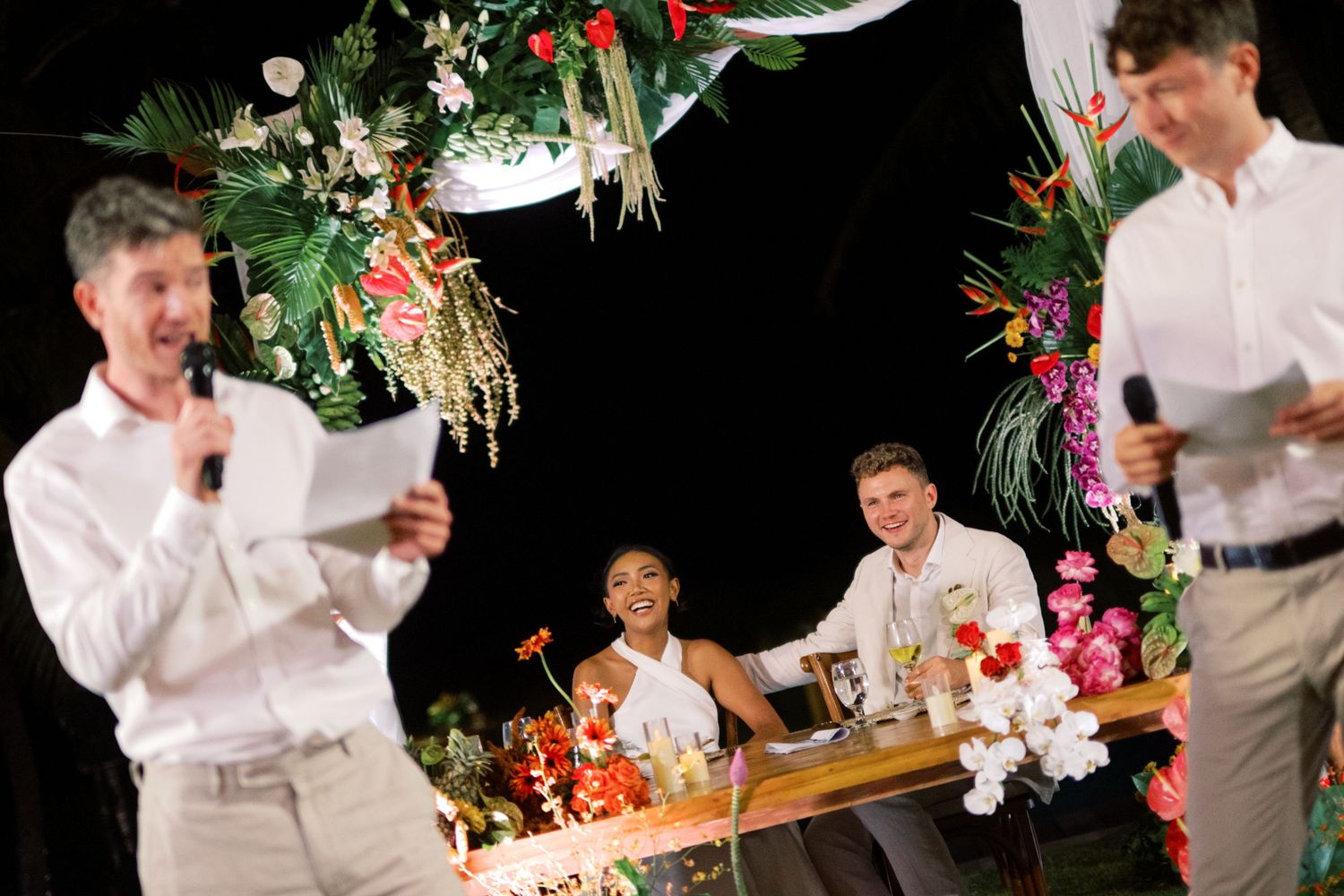Two men give speeches at a tropical wedding reception while the bride and groom smile at a flower-adorned table.
