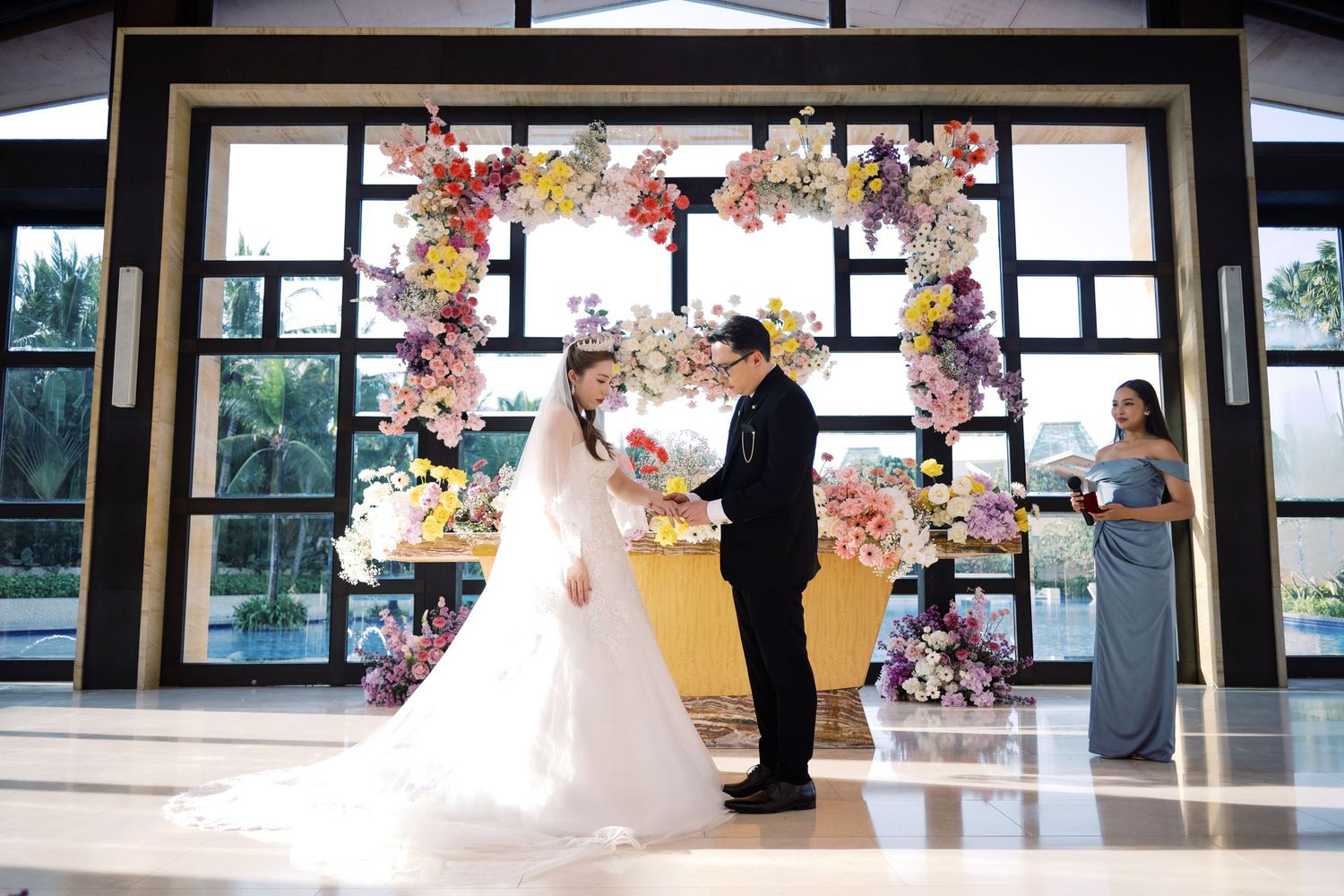 Couple exchanging vows at elegant wedding ceremony with colorful floral arch backdrop and bridesmaid in gray dress.