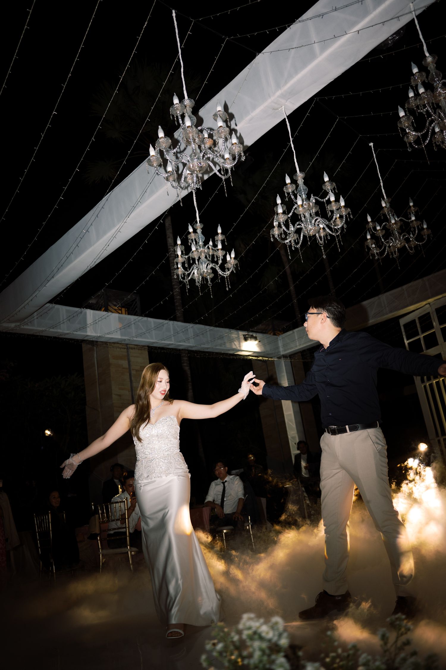 Bride and groom share first dance under crystal chandeliers with dramatic fog effects at elegant wedding reception.