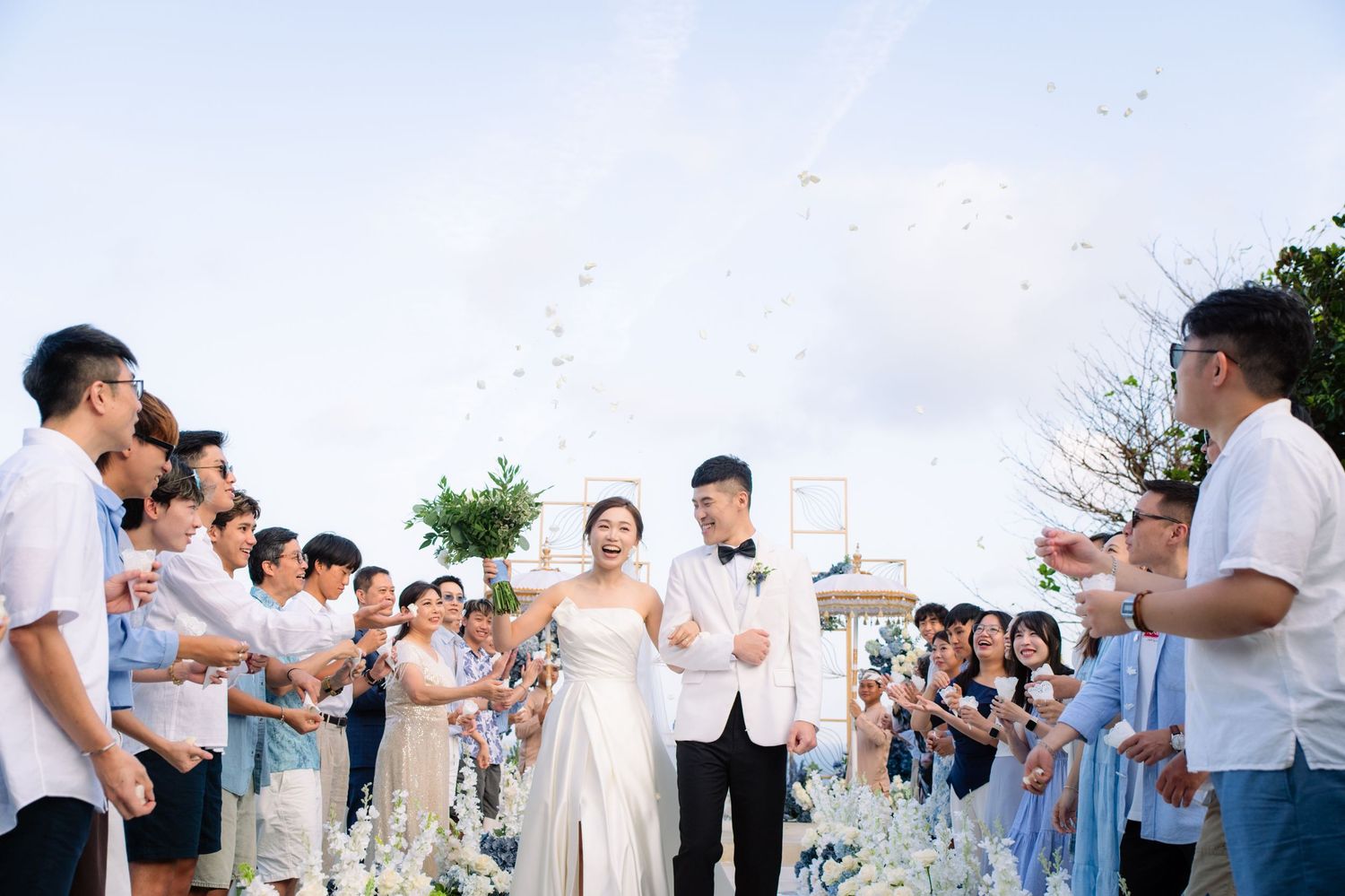 Bride and groom walk down a beachside wedding aisle as smiling guests toss petals and cheer on both sides under a bright blue sky.