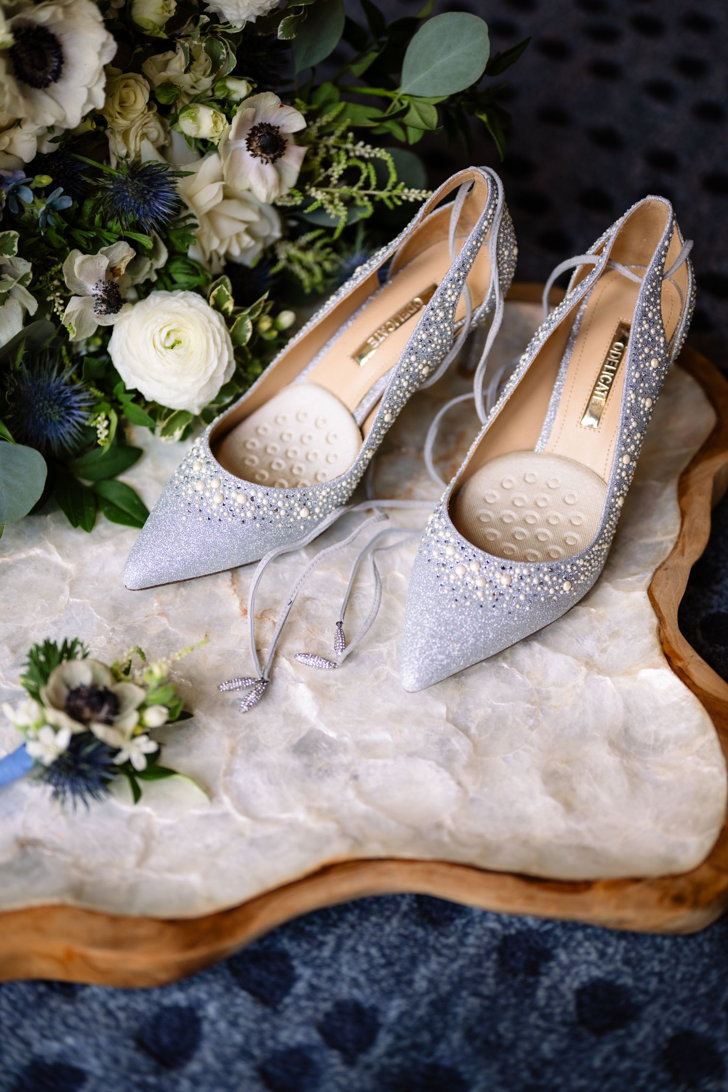 Sparkling silver glitter bridal heels with ankle ties on a marble tray beside a bouquet of white roses and blue thistle.