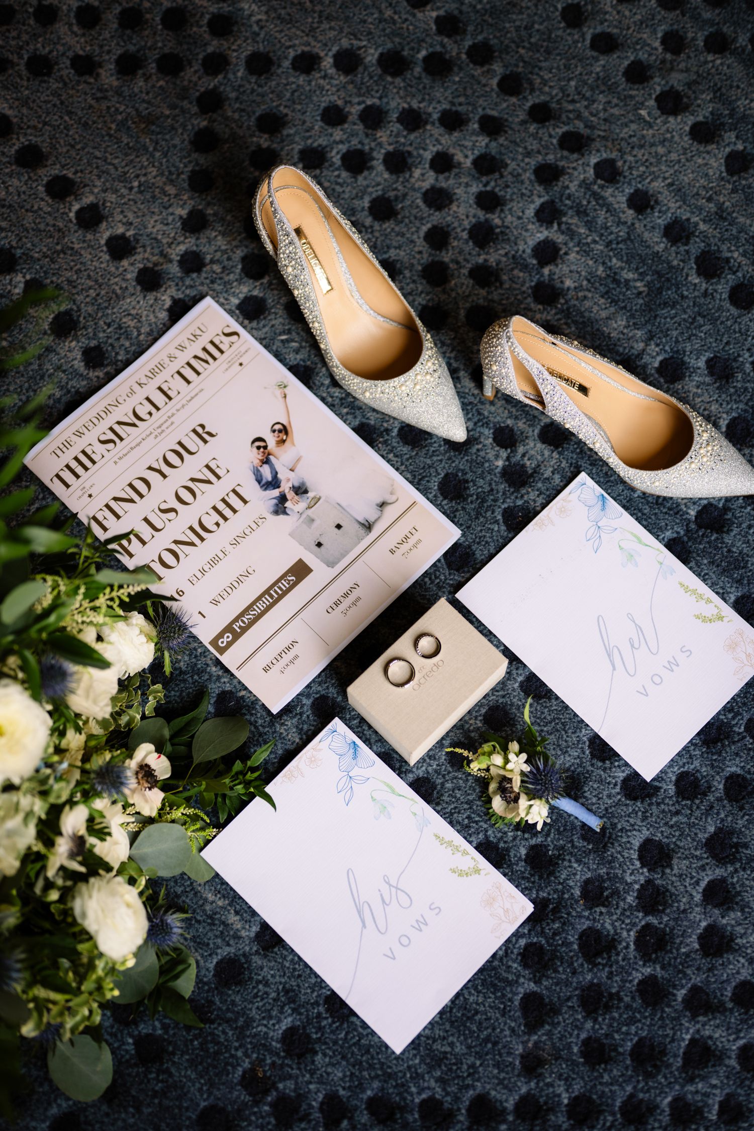 Wedding flat lay with glittery heels, his and hers vow booklets, a ring box, a boutonniere, white flowers and a newspaper-style program.