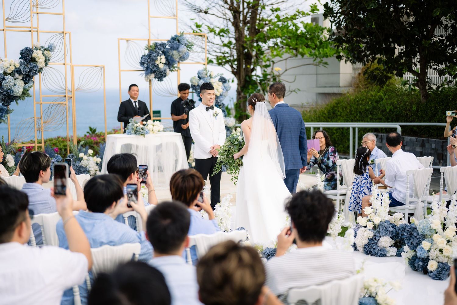 Bride and groom stand before an officiant during an outdoor seaside wedding ceremony as guests photograph the moment with their phones.