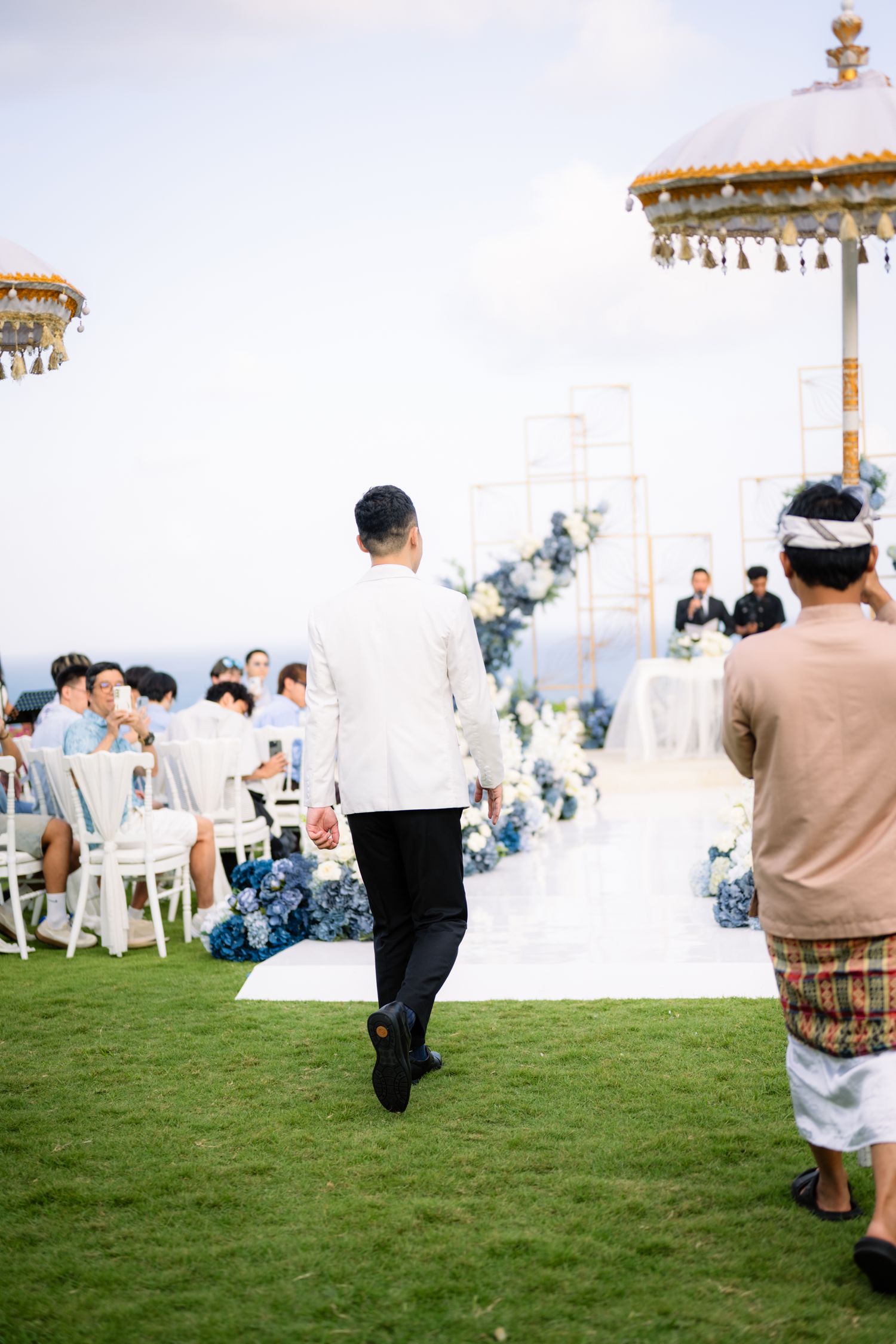 Groom in a white jacket walks toward a seaside wedding ceremony with blue floral arrangements, guests seated, and ornate umbrellas.