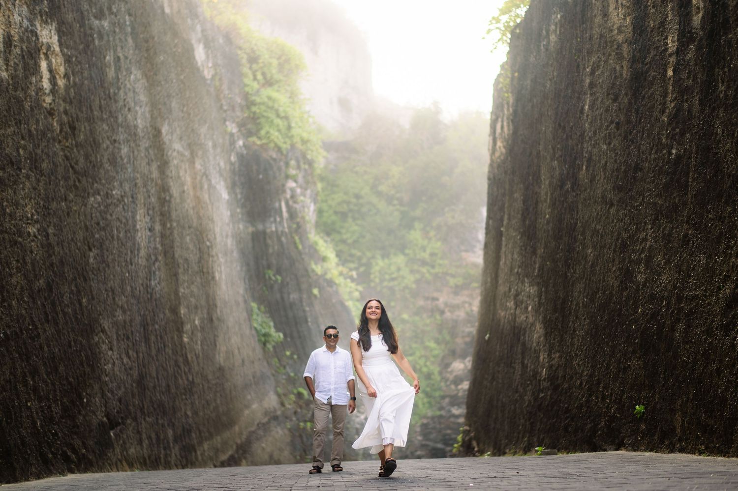 Couple in white outfits walking through a dramatic rocky canyon passage with misty green cliffs in the background.