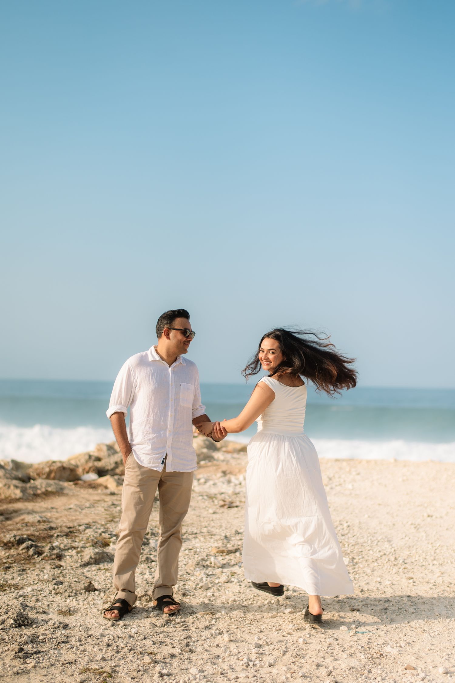 Couple holding hands and walking on a sunny beach, woman in white dress with hair blowing, man in white shirt and khaki pants.
